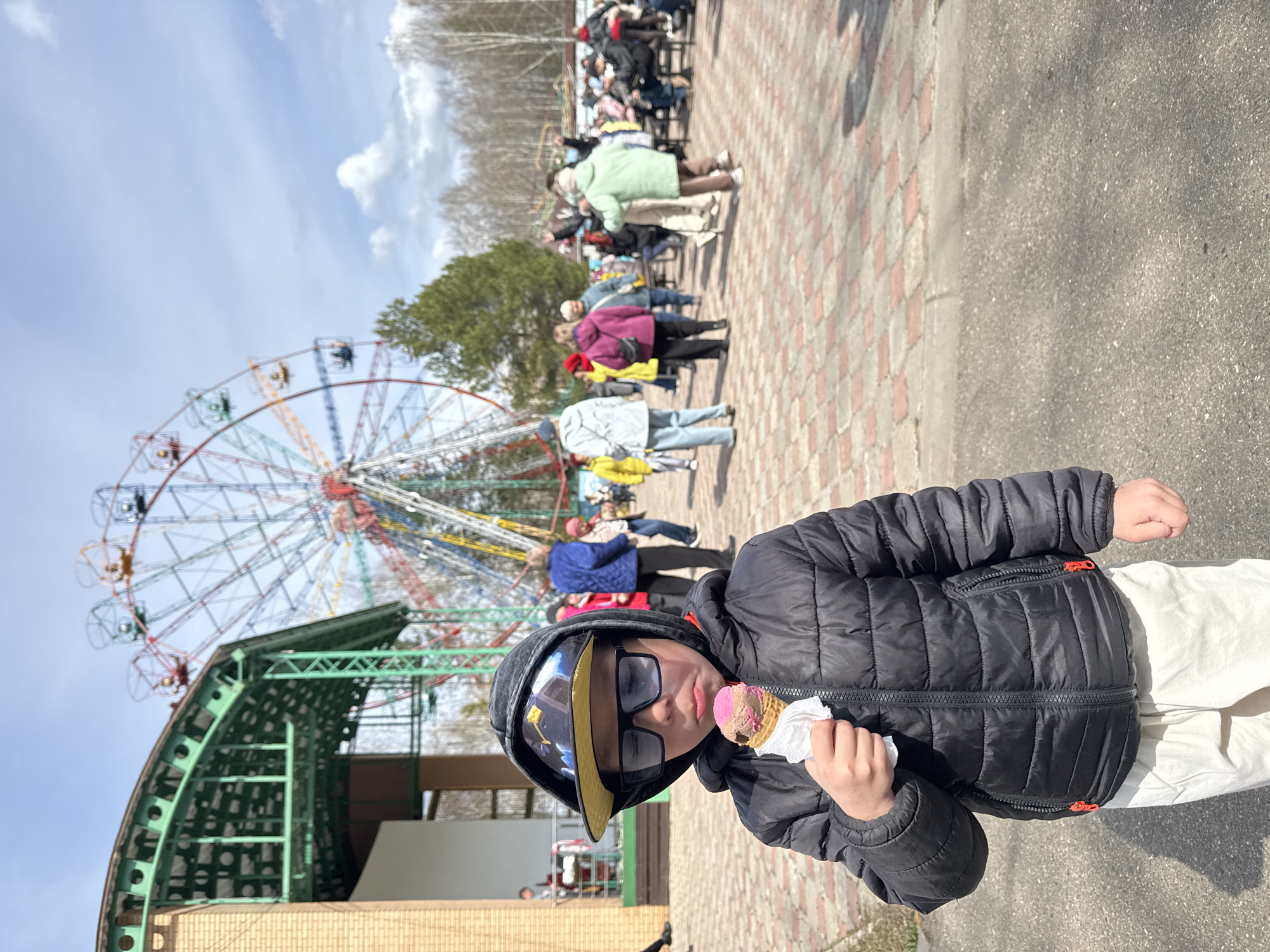 This image captures a bright, sunny day at what appears to be an amusement park or fairground in Chaikovsky, Russia. The primary subject is a young boy, approximately 6-10 years old, positioned prominently in the foreground. He is wearing a black hooded puffer jacket, dark rectangular sunglasses, and a black baseball cap with a striking yellow brim. The boy is holding a two-scoop ice cream cone, featuring pink and brown swirls, wrapped in a white napkin, and appears to be enjoying it.
In the mid-ground, a large, multi-colored Ferris wheel with several cabins is a dominant feature, set against a clear blue sky dotted with scattered white clouds. Below the Ferris wheel, a distinctive green, arched architectural structure is visible, likely part of the ride's base or an adjacent building.
To the right and further into the background, several adults and children are visible, walking along paved pathways made of patterned brick-like tiles and asphalt. They are dressed in various casual jackets and coats, indicating a cool but pleasant temperature. Bare trees in the distance suggest either early spring or late autumn. The scene depicts a lively, leisurely atmosphere typical of a public park or fair. No visible text is discernible in the image.