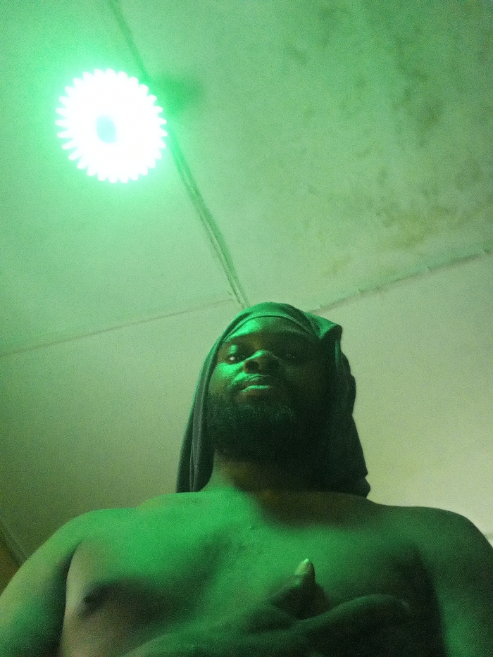 The image features a man illuminated by a bright green light from above, taken from a low-angle perspective looking upwards. The man has dark skin and a beard, and a dark piece of fabric is draped over his head, possibly a head covering or part of his attire. He is shirtless, and the green light strongly bathes his chest and face, creating highlights and shadows. His eyes are open, and he appears to be looking directly into the camera or slightly upwards towards the light source.
In the upper part of the frame, a distinct, bright circular light fixture is visible on the ceiling. This fixture emits a strong white light from its center, but the overall illumination in the scene is a vibrant green, suggesting the light source itself is green, or a green filter is in place. The light fixture has a serrated or spiky edge, resembling a gear or cog. Wires are visible on the ceiling, leading towards this light.
The ceiling itself shows signs of wear or possible water damage, with visible discoloration and stains in several areas, particularly in the upper right portion of the image. The overall setting appears to be indoors, likely a room with a plain ceiling. The intense green light creates a distinct, almost otherworldly or dramatic atmosphere for the portrait. The man's left hand is visible in the lower right corner, with fingers partially clenched, though the exact gesture or what he might be holding is unclear due to the lighting and angle. No text is visible in the image.