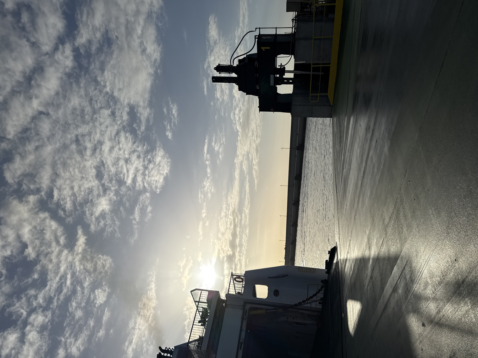 The image depicts a scene on a vessel in Yaiza, Spain, potentially a ferry or a large boat, at dawn or dusk given the low sun and cloudy sky. To the right of the frame is a section of the vessel's deck, appearing wet or polished. A piece of heavy machinery is visible, possibly part of the vessel's equipment. A coastline is discernible in the distance with what appears to be buildings or structures lined along the coast. To the left are white cumulus clouds. In the foreground are sections of the vessel, including railings and life-saving equipment.