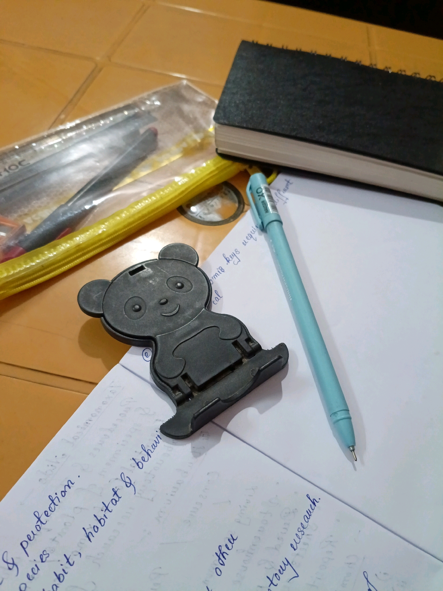The image depicts a cluttered desk surface with various study-related items. In the foreground, a black panda-shaped phone stand is prominently displayed. To the right of the stand, a light blue pen lies on a sheet of paper filled with handwritten text. The paper appears to be part of a notebook, with some lines visible. Behind the pen and paper, a black spiral-bound notebook is partially visible. To the left of the panda stand, a yellow pencil case is open, revealing some stationery items inside, including what appear to be pens or pencils. The background is a wooden surface, possibly a table or desk. There are no people visible in the image. The lighting suggests an indoor setting, likely during the daytime. The scene is consistent with a study area or workspace. The image does not contain any information that can determine its location in Bhatpara, India.