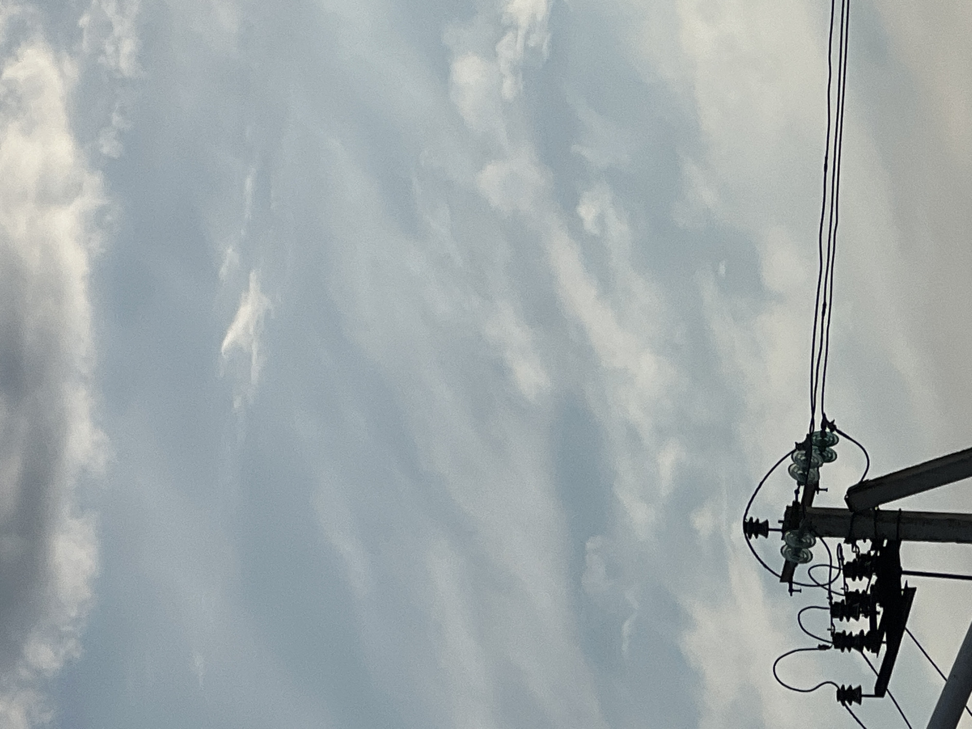 An outdoor, medium shot captures a utility pole with electrical equipment against a cloudy sky in Golubitskaya, Russia. The pole, made of concrete, supports multiple insulators and power lines. The sky is filled with scattered clouds, some appearing wispy and others denser, with a pale blue hue visible in between. The scene is devoid of any people or animals. The focus is on the infrastructure against the natural backdrop of the sky.