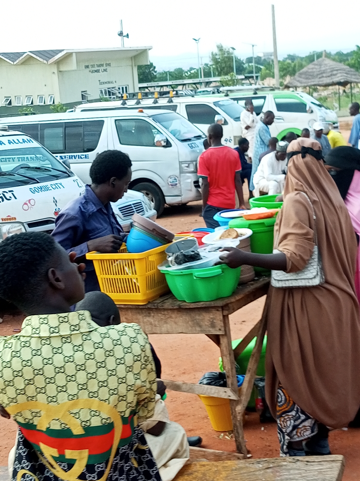 The image depicts an outdoor scene at a transportation terminal in Gombe City, Nigeria. In the background, a building labeled "GOMBE LINE TERMINAL 4" is visible, along with several white minivans. The foreground features a wooden stall laden with various food items served in plastic bowls and plates. Several people are gathered around the stall, some appearing to be customers and others vendors. A woman in a brown hijab is interacting with the stall, reaching for a plate. Another person, wearing a blue shirt, is handling plastic containers. The ground is reddish-brown, suggesting a dusty or sandy environment. The overall atmosphere appears to be that of a busy public transportation hub with food vendors serving travelers.