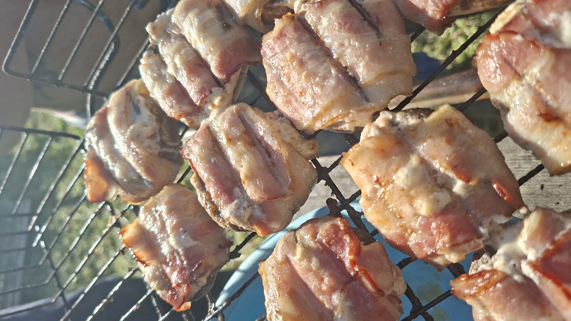 The image depicts several pieces of bacon cooking on a metal grill. The bacon is arranged in a grid pattern and appears to be in the process of being cooked. The environment visible behind the grill suggests an outdoor setting, possibly a backyard or picnic area. The location is Сваткаўскі сельскі Савет, Belarus. No people are visible in the image.