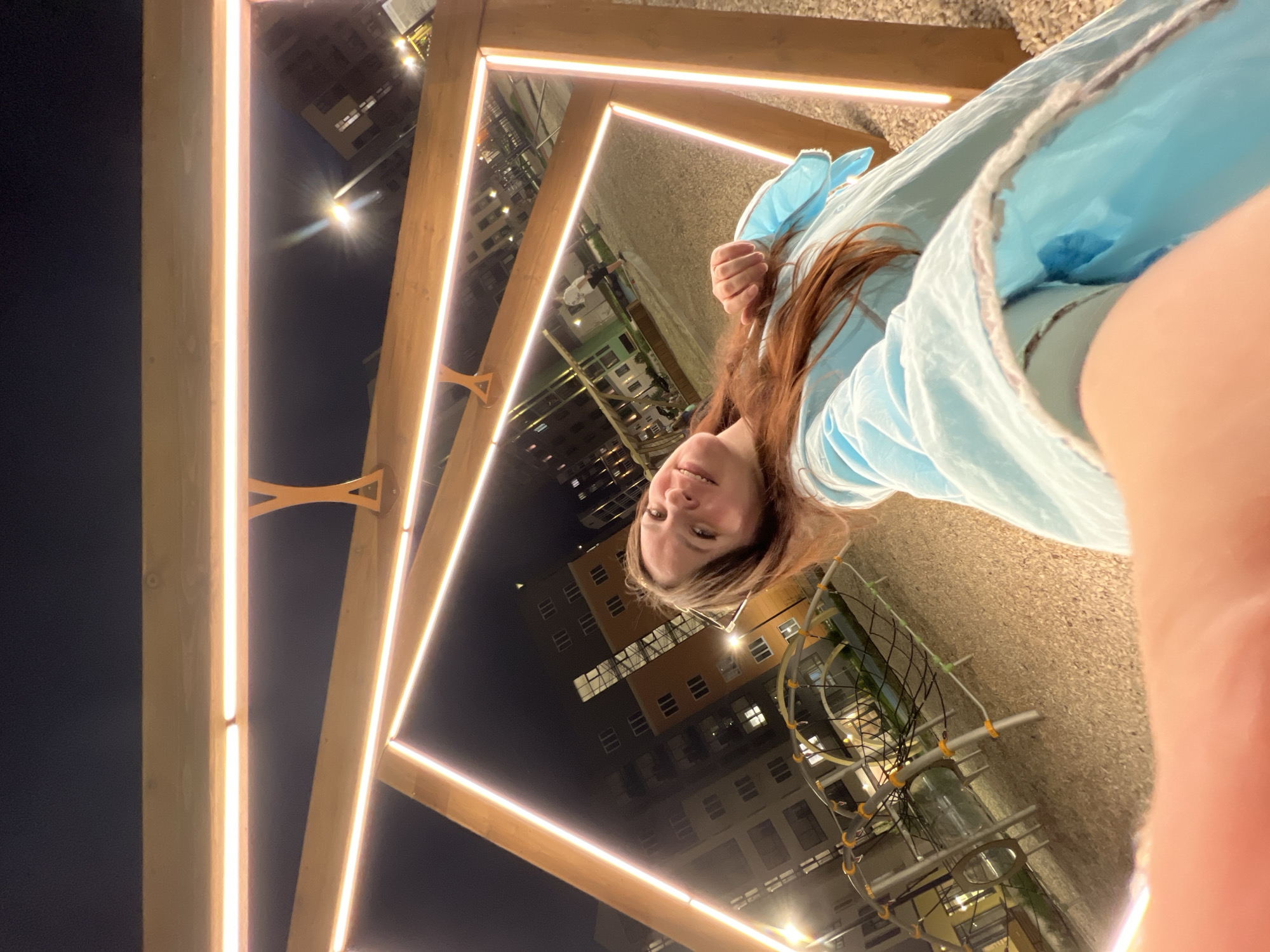 A selfie taken at night in Pervomaysky, Russia, shows a woman with reddish-brown hair and a light blue jacket. She is positioned within a modern art installation of interconnected wooden frames illuminated with LED strip lights. Buildings with lighted windows are visible in the background, as is a playground structure with metal bars and netting.
