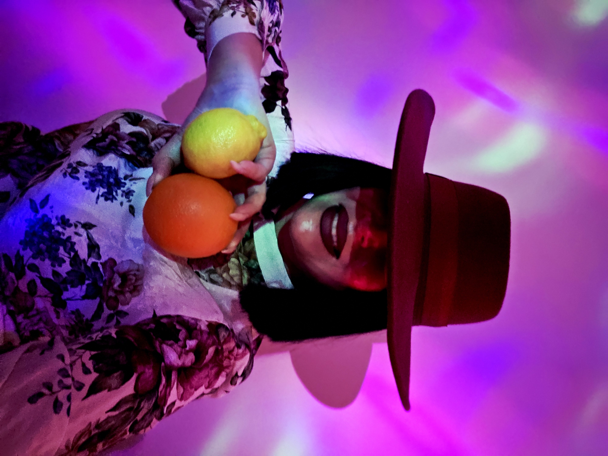 The image captures a person from an overhead perspective, rotated 90 degrees counter-clockwise so that their head is on the right side of the frame and their body extends leftwards. The individual has dark hair and is wearing a wide-brimmed reddish-brown hat. Their face is partially obscured by the hat's brim and shadows, but their mouth is open, revealing teeth, suggesting a smile or speaking. They are holding a bright yellow lemon in their right hand and a vibrant orange in their left, both prominent in the foreground.
The person is dressed in a garment with a floral pattern, featuring dark blue or purple flowers and foliage on a lighter background, although the colors are heavily influenced by the ambient lighting. The background is dominated by intense purple and pink hues, with blurred lighter patches of magenta and light green/white, indicating a dynamic, artificial lighting environment, possibly from a disco light or colorful spotlights. No specific location cues like "indoors" or "outdoors" are discernible, but the lighting suggests an indoor setting or nighttime. The overall mood is vibrant and energetic due to the strong, artificial colors. There is no visible text in the image.