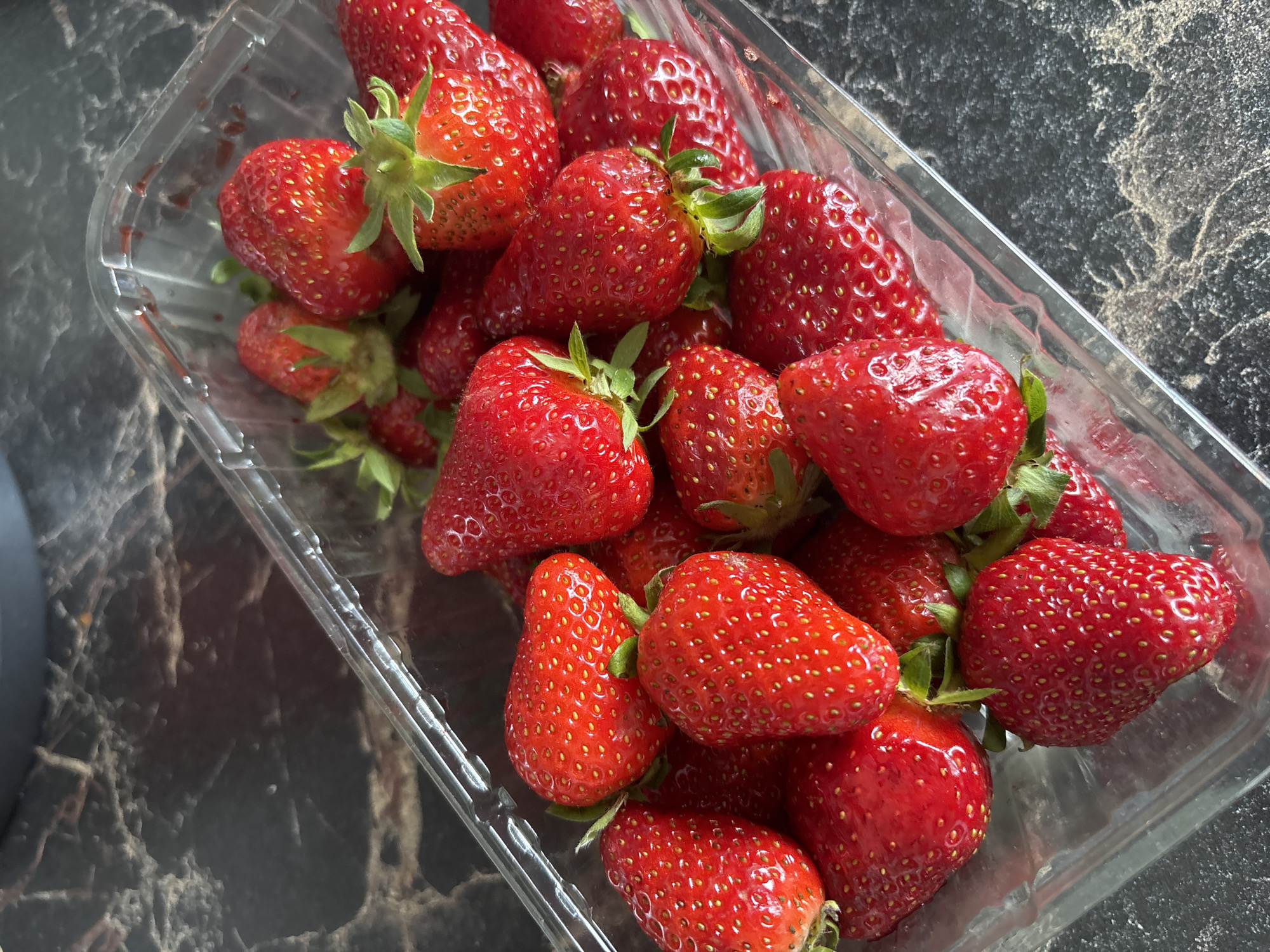 A clear, plastic container is filled with fresh, ripe strawberries. The strawberries are a vibrant red color and have visible seeds on their surface. Some have green stems still attached. The container sits on a dark, marble-like surface. The image contains no indication of being located in Sochi, Russia.