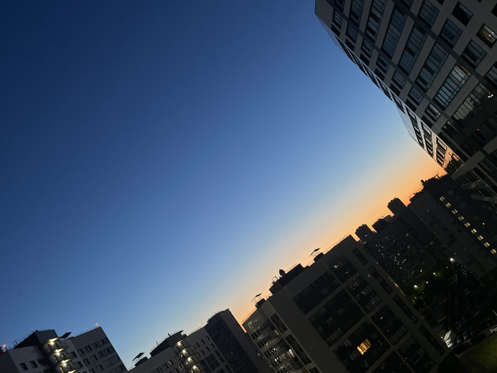 An angled, exterior, low-light, full shot captures a twilight sky above a dense urban landscape in Sinyavino, Russia. The sky transitions from a deep sapphire blue at the top of the frame to a soft, pale orange near the horizon, indicating sunset or sunrise. Silhouetted against the sky are numerous apartment buildings of varying heights and architectural styles. Some buildings exhibit visible window lights, suggesting occupancy. The buildings are arranged in rows and at different angles, creating a sense of depth and urban density. The overall atmosphere is serene, with the city lights beginning to twinkle as the natural light fades. There are no people or vehicles prominently visible in the frame.