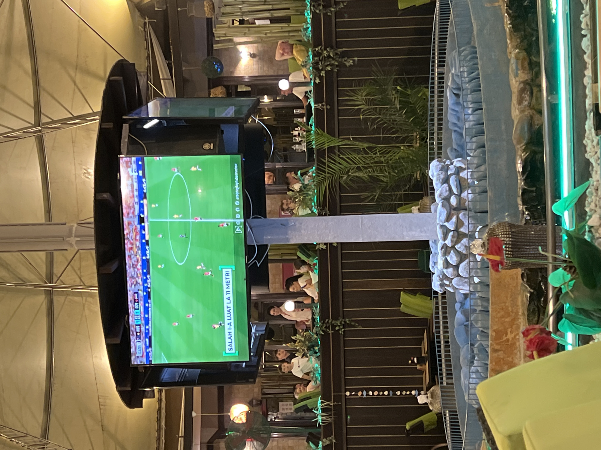 The image captures an indoor scene at a venue in Constanța, Romania, likely a bar or restaurant, where a football match is being displayed on a large screen. The screen shows a soccer game in progress, with players on a green field. Below the screen, text in what appears to be Indonesian or Malay reads "SALAH 1-A LUAT LA 11 METRI," which translates to "Salah takes the 11-meter penalty."
Several groups of people are visible seated at tables and railings, watching the screen. In the foreground, a decorative fountain-like structure with stones and some greenery is present. The overall ambiance suggests a casual setting for patrons to watch sports. The lighting is warm and ambient, with some artificial lights visible throughout the space.