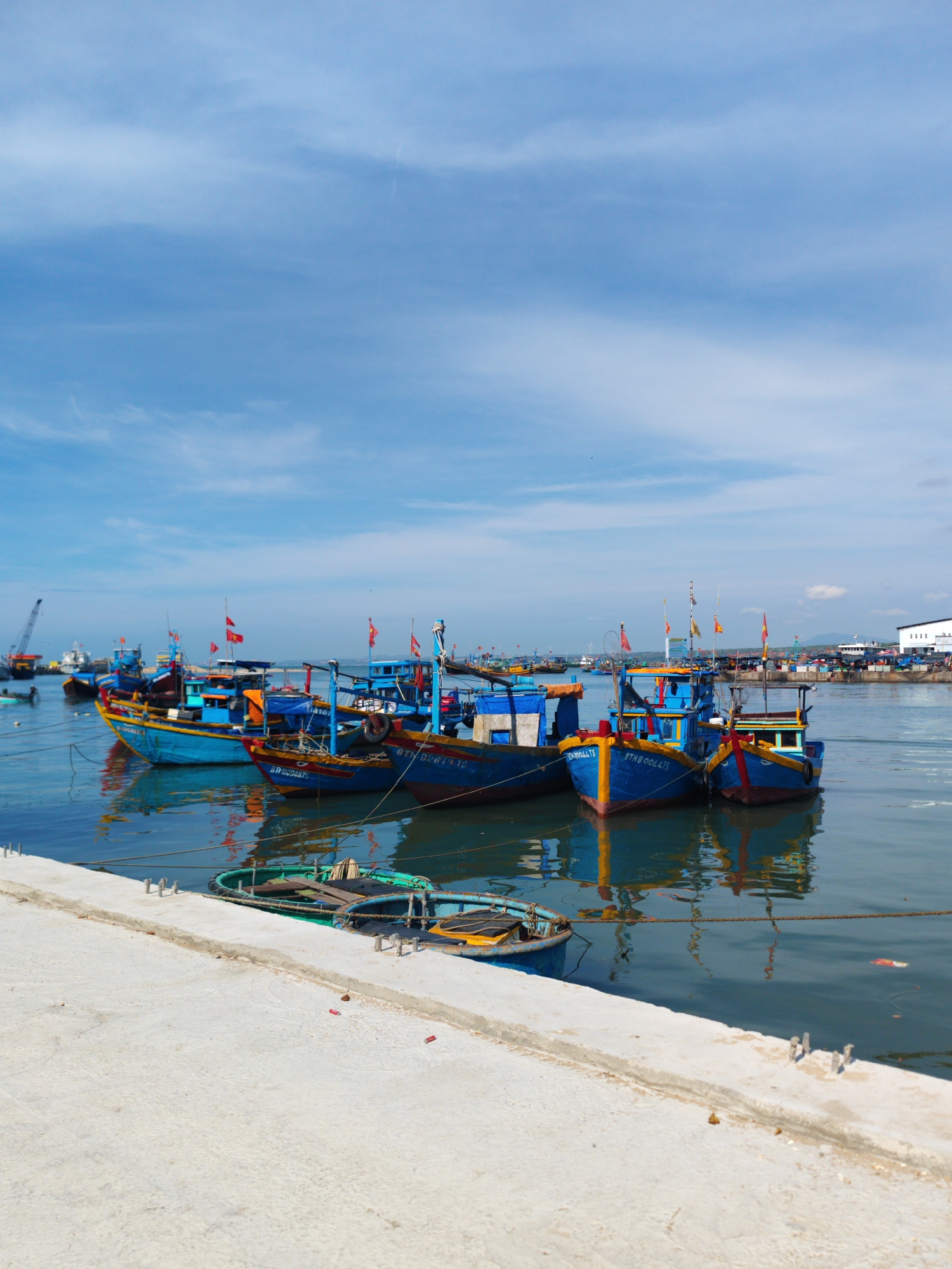 The image shows several blue and yellow fishing boats docked along a concrete pier in the city of Phan Thiết, Vietnam. The boats are tied together and to the pier with ropes. Each boat has a mast with small flags. The water in the harbor is calm, reflecting the boats and the clear blue sky with scattered clouds. In the background, there is a line of buildings and other boats, suggesting a busy port area. The pier is light gray and appears to be freshly constructed or well-maintained. There is some debris floating in the water near the pier. A small green rowboat is partially visible in the foreground, next to the pier.