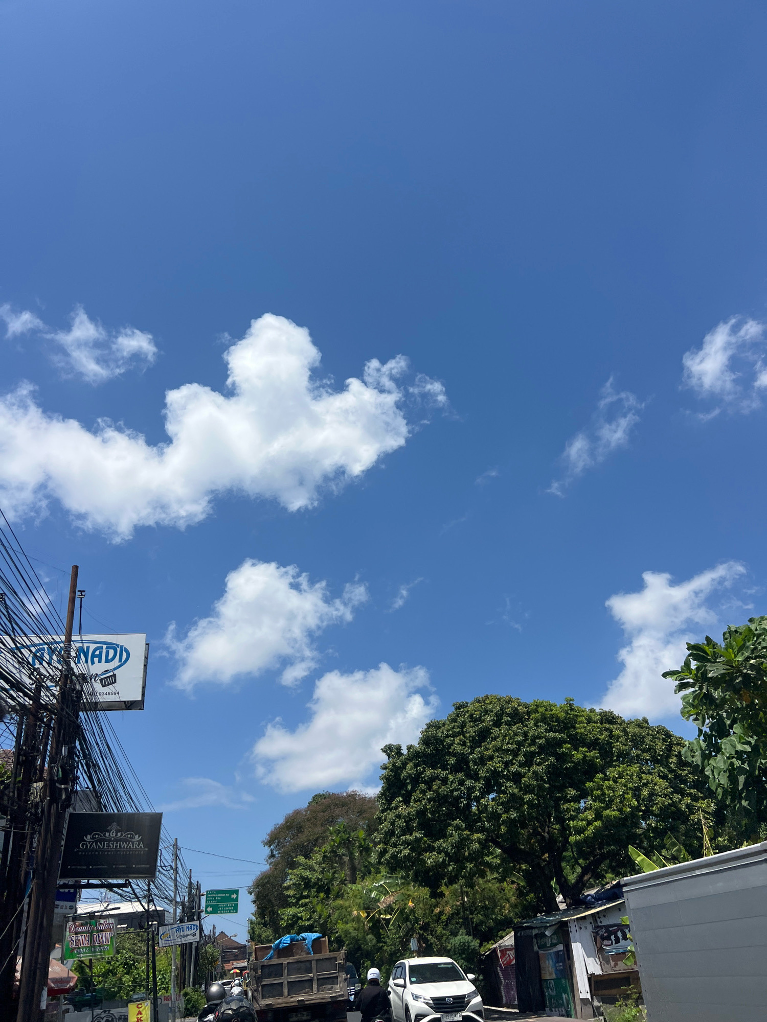 The image captures a street scene in Denpasar, Indonesia, under a clear blue sky dotted with scattered white clouds. A dense network of electrical wires dominates the left side of the frame, originating from a wooden pole and crisscrossing the sky. Below the wires, several signs are visible. One sign, partially obscured, reads "ATENADI" with a phone number. Another, larger sign displays "GYANESHWARA" in prominent lettering, followed by smaller text, suggesting a business or establishment. Further down the street, more signs and traffic can be observed.
In the foreground, a white car is parked on the right side of the road, and a truck with a load covered in blue tarpaulin is also present. Several motorcycles with riders are navigating the street. Lush green trees, including a large, mature tree with a full canopy, form a backdrop to the scene, interspersed with smaller structures and buildings. The overall impression is one of a busy urban environment on a sunny day.