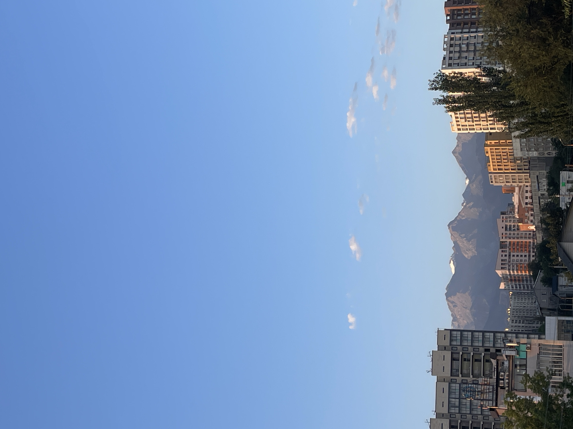 The image captures a cityscape against a backdrop of majestic mountains under a clear blue sky dotted with a few wispy clouds. In the foreground, various multi-story buildings, characteristic of Almaty, Kazakhstan, are visible. These structures exhibit diverse architectural styles, from modern apartment complexes with balconies and windows to older buildings with more traditional facades. Lush green trees punctuate the urban landscape, adding a touch of nature to the scene.
The mountains, with their snow-capped peaks, dominate the horizon, suggesting a proximity to a range like the Tian Shan mountains, which famously surround Almaty. The sunlight illuminates the buildings, casting warm hues on some of the facades, indicating either sunrise or sunset. The overall atmosphere is tranquil and picturesque, showcasing the harmonious coexistence of urban development and natural grandeur. No people or specific actions are discernible in the image, suggesting a static scene captured from a vantage point overlooking the city.