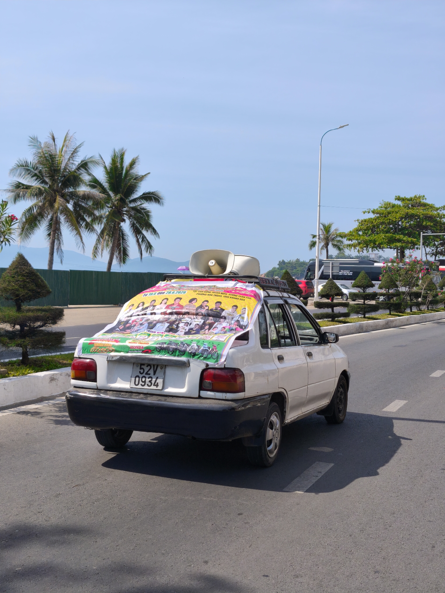 The image shows the rear and side of a white sedan in Bac Nha Trang Ward, Vietnam. The car is on an asphalt road with white lane markings. A large banner is draped over the rear of the car, featuring a group of people and several motorcycles. The banner also has text in Vietnamese, including "Tu 19.5 den 26.6.2026" and what appears to be a street address or location. On the roof of the car, a loudspeaker is mounted. In the background, there are palm trees, a green fence, a street light, and other vehicles, including a bus. The sky is clear and blue. The license plate of the car reads "52V 0934".