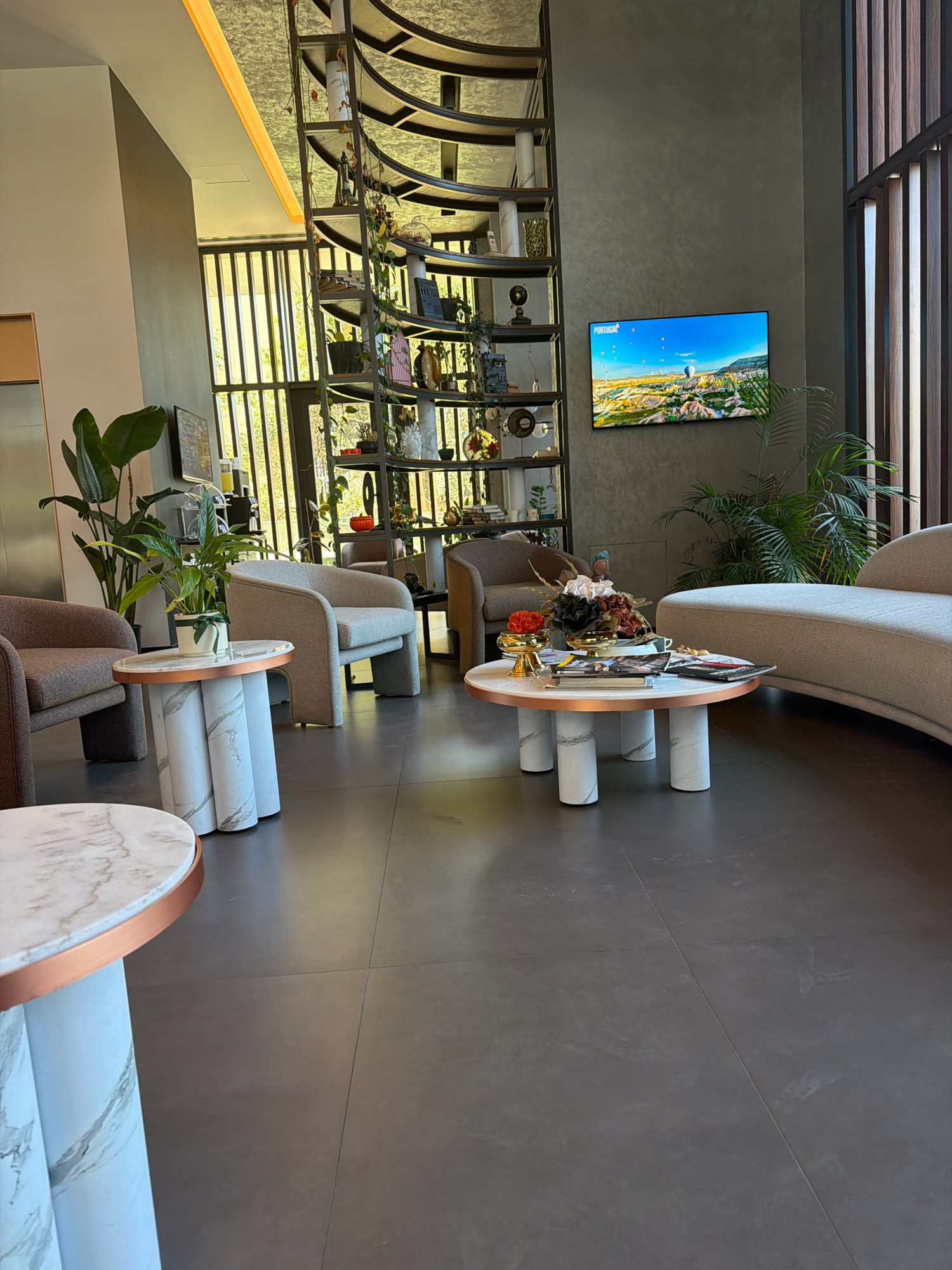 This image depicts an indoor seating area, likely a hotel lobby or lounge, in Bakırköy, Turkey. The scene is furnished with modern, minimalist decor. In the foreground, a round coffee table with a marble-patterned top and cylindrical legs with a copper trim is visible. Next to it, a smaller, similarly designed side table holds a potted plant.
Behind these tables, several upholstered armchairs are arranged. One is a light grey, curved armchair, and another is a deeper brown armchair. Further back, a long, curved sofa with a light beige upholstery dominates the right side of the frame. A large, multi-tiered metal shelving unit, filled with various decorative items, plants, and what appear to be books, stands prominently against a textured grey wall on the left.
A flat-screen television mounted on the wall displays an image of hot air balloons over a landscape, with the word "PORTUGAL" visible in the top left corner. Large windows with vertical wooden slats allow natural light to enter the space. A tall houseplant is positioned to the left of the television. The floor is tiled with large, dark grey tiles. The overall ambiance is one of contemporary elegance and comfort.