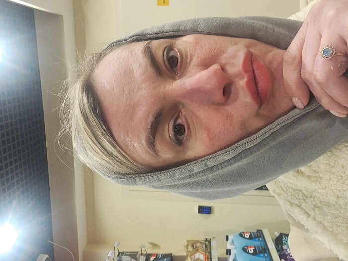 A fair-skinned woman with light blonde hair and a gray hoodie is captured in a close-up shot. She has brown eyes and red lips. Her right hand is visible, displaying a ring with a blue stone. The background reveals a wall, shelves with products, and a portion of a window or screen reflecting light.