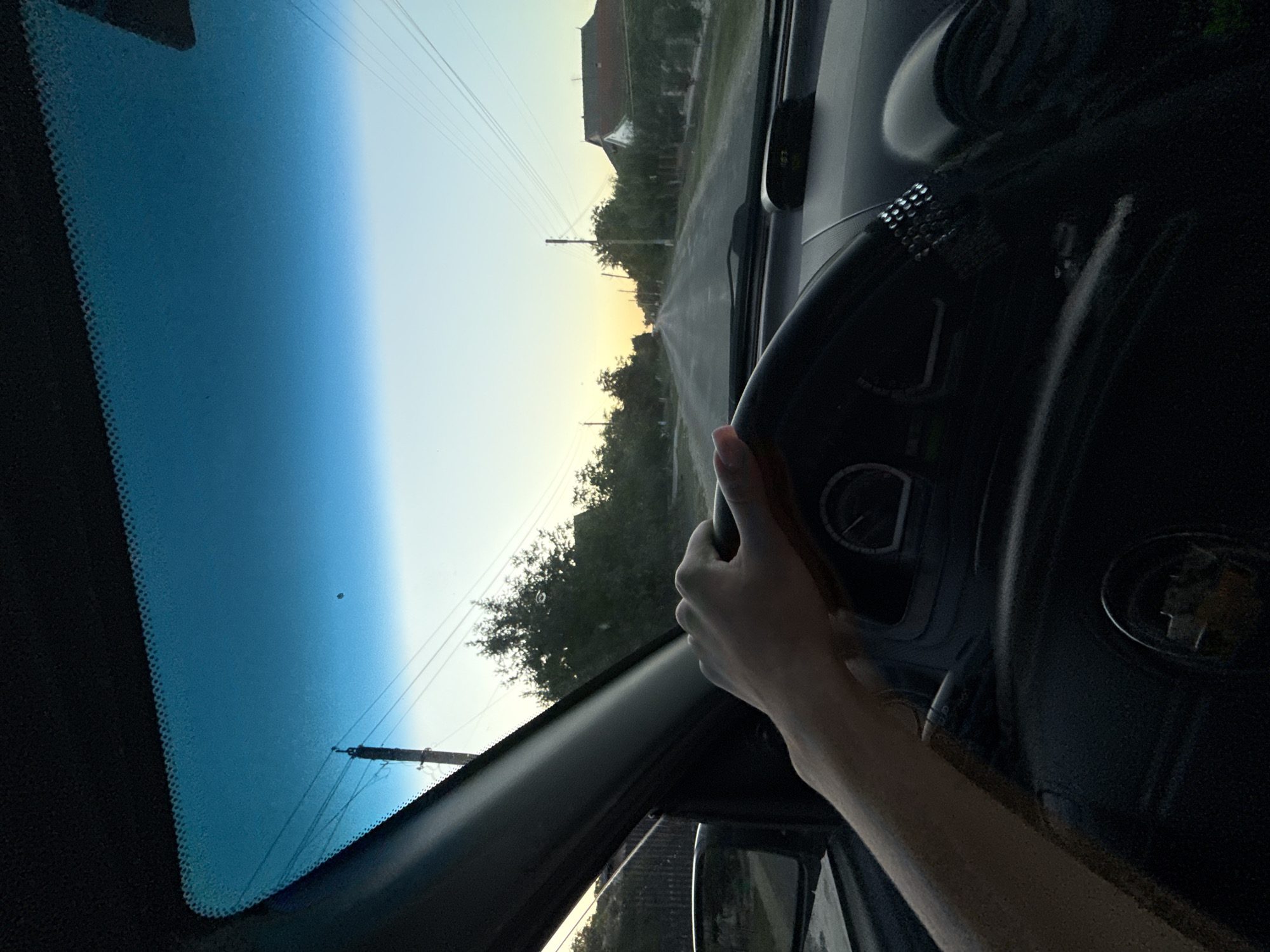 An interior shot from the driver's perspective shows a person's left hand resting on a black steering wheel. The hand is light-skinned and appears to be male. Through the windshield, a paved road stretches into the distance, lined with trees and houses. The sky is a pale yellow and blue, suggesting either sunrise or sunset. A utility pole with power lines is visible on the left side of the road. The scene is set in a rural or suburban area, likely in Cioc-Maidan, Moldova, given the context. The overall atmosphere is calm and ordinary, depicting a drive during twilight.