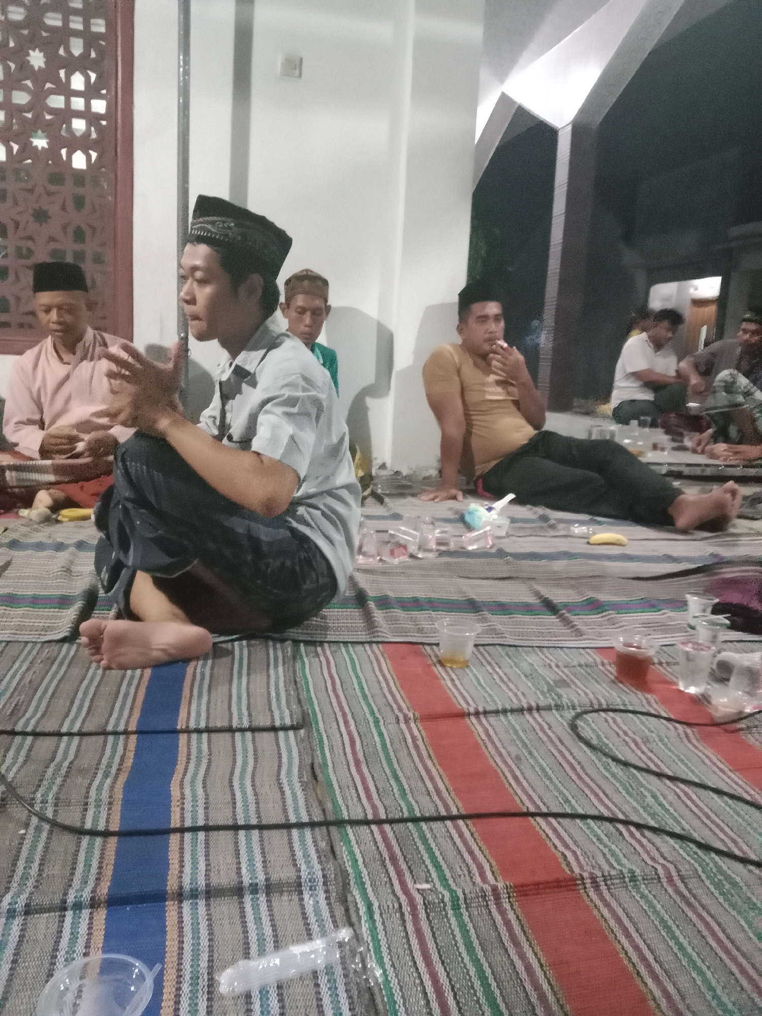 In Brangsong, Indonesia, a group of men are gathered indoors, seated on a striped mat. A man in the foreground wears a black cap and a light-colored collared shirt, with his hands clasped in front of him. To his left, another man in a pink shirt and black cap sits with his hands on his lap. Behind them, a man in a teal shirt is visible. Further back, a man in a yellow t-shirt leans against a wall, smoking. Other men are seated in the background. Various plastic cups and a banana are scattered on the mat, suggesting a casual gathering. The environment appears to be a room with white walls and a visible window with ornate latticework on the left.