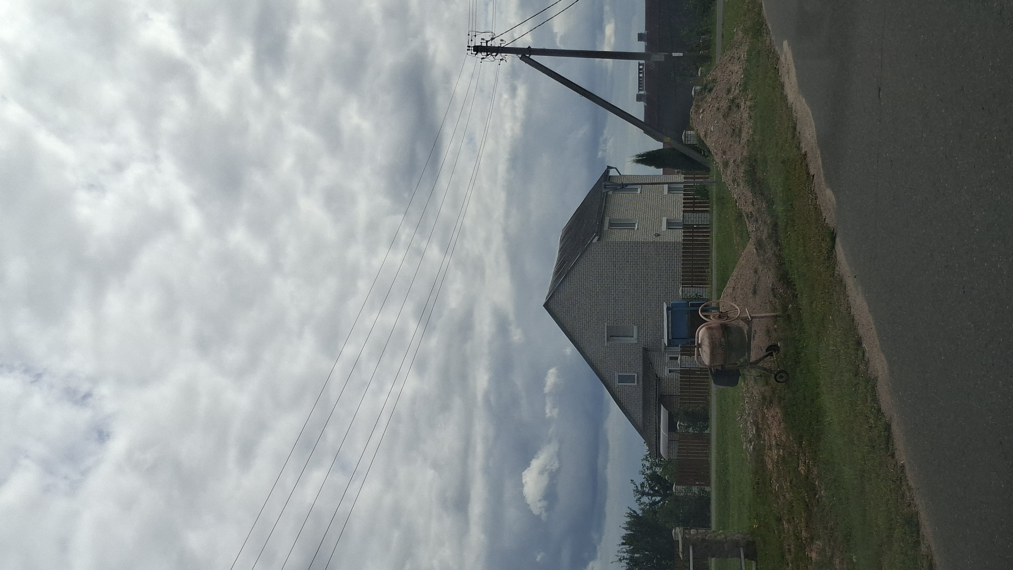 The image depicts a residential scene on a cloudy day in Myadzyel, Belarus. In the foreground, a paved road runs diagonally from bottom right to the left. To the left of the road, a patch of dry grass and a concrete mixer with a pile of dirt behind it are visible. Further left, a wooden fence delineates the property line.
Dominating the mid-ground is a two-story house constructed of light-colored brick, with a dark, sloped roof. The house has multiple windows on both levels. A small, covered porch is attached to the front left of the house. Adjacent to the house, a blue portable toilet is situated.
In the background, a wooden utility pole with electrical wires stretching across the sky stands prominently on the right. Several other houses and trees are visible in the distance, extending towards the horizon. The sky is heavily overcast with gray and white clouds. No people are present in the image.