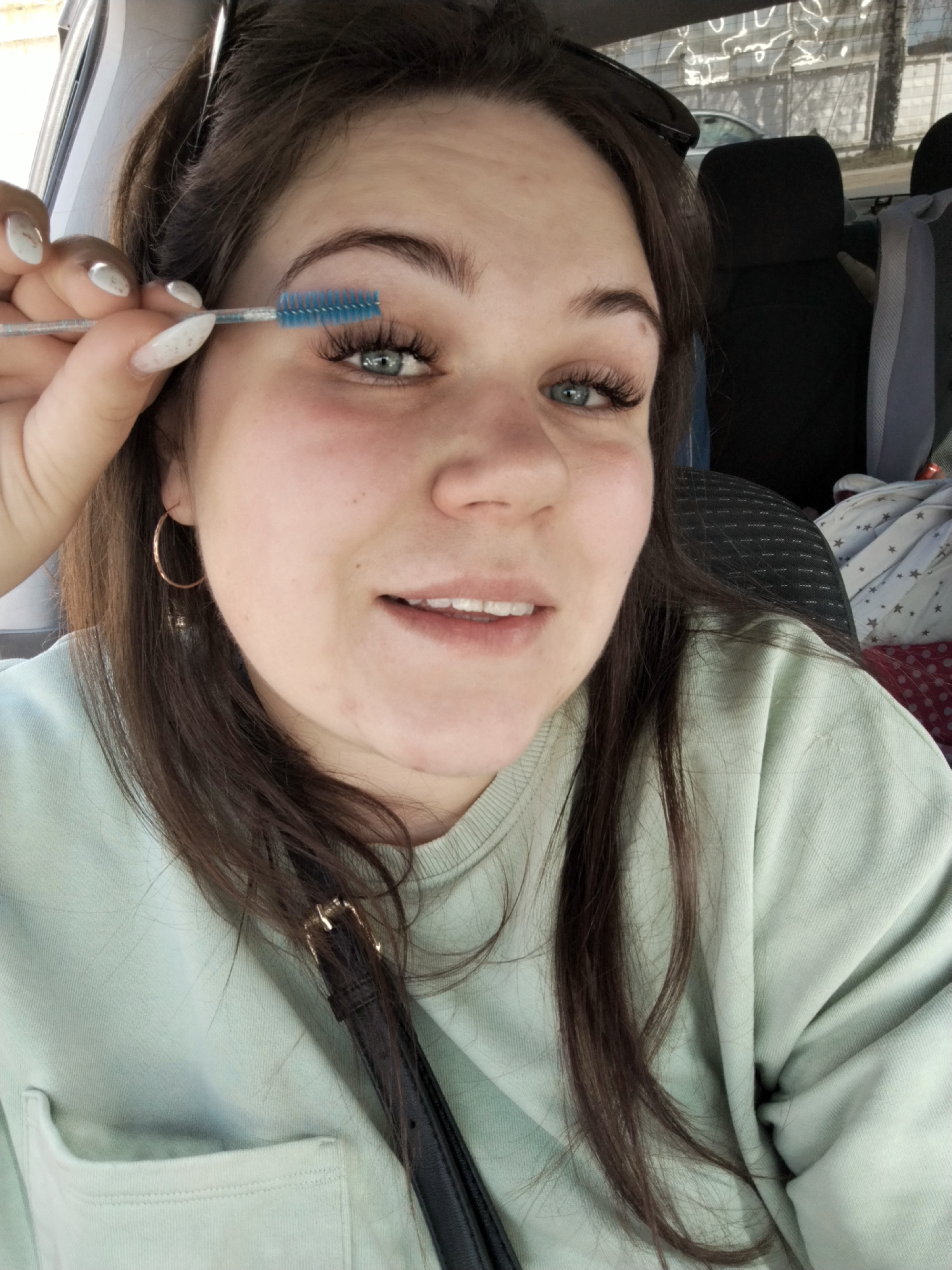 A young woman is inside a vehicle. She is fair-skinned with brown hair and blue eyes. She is wearing a light green sweatshirt and gold hoop earrings. Her fingernails are long and painted white with glitter accents. She is holding a blue eyelash brush near her right eye. The vehicle's interior shows black headrests, seats, and a blanket with white stars. The location is Chaikovsky, Russia.