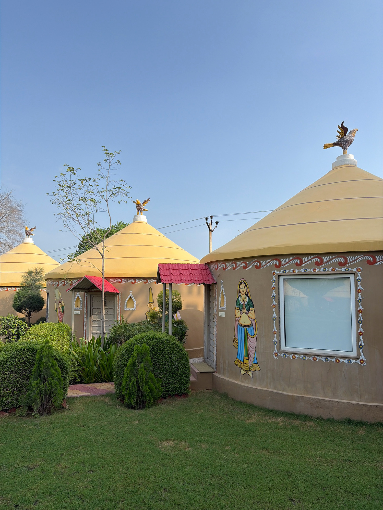 The image shows a row of circular, tan-colored huts with conical yellow roofs in Dockiya, India. Each hut is decorated with traditional Indian art, including paintings of women in colorful attire and floral borders. Bird figurines are perched on top of the roofs. The huts are surrounded by lush green grass and manicured bushes. The sky is clear and blue. The scene appears to be a tourist resort or a cultural village.