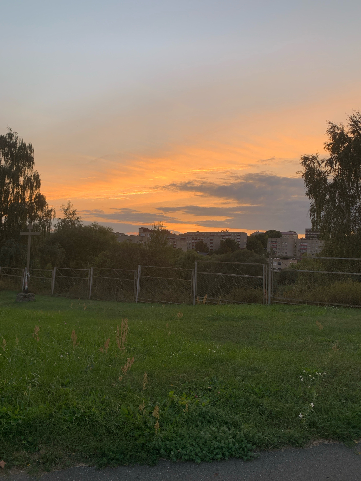 The image depicts an outdoor scene at dusk in the city of Kapyĺ, Belarus. The sky is filled with a vibrant orange and yellow sunset, with scattered clouds. In the foreground, a grassy field extends across the bottom half of the frame. A chain-link fence runs horizontally through the middle ground, separating the field from a line of trees and buildings. To the left, a wooden cross stands in the field, with a small statue at its base. Behind the fence, dense foliage obscures the lower portions of several apartment buildings, which appear to be Soviet-era construction. A large tree with dark foliage is visible on the far right. The overall impression is one of a quiet, somewhat bleak urban landscape at the end of the day.