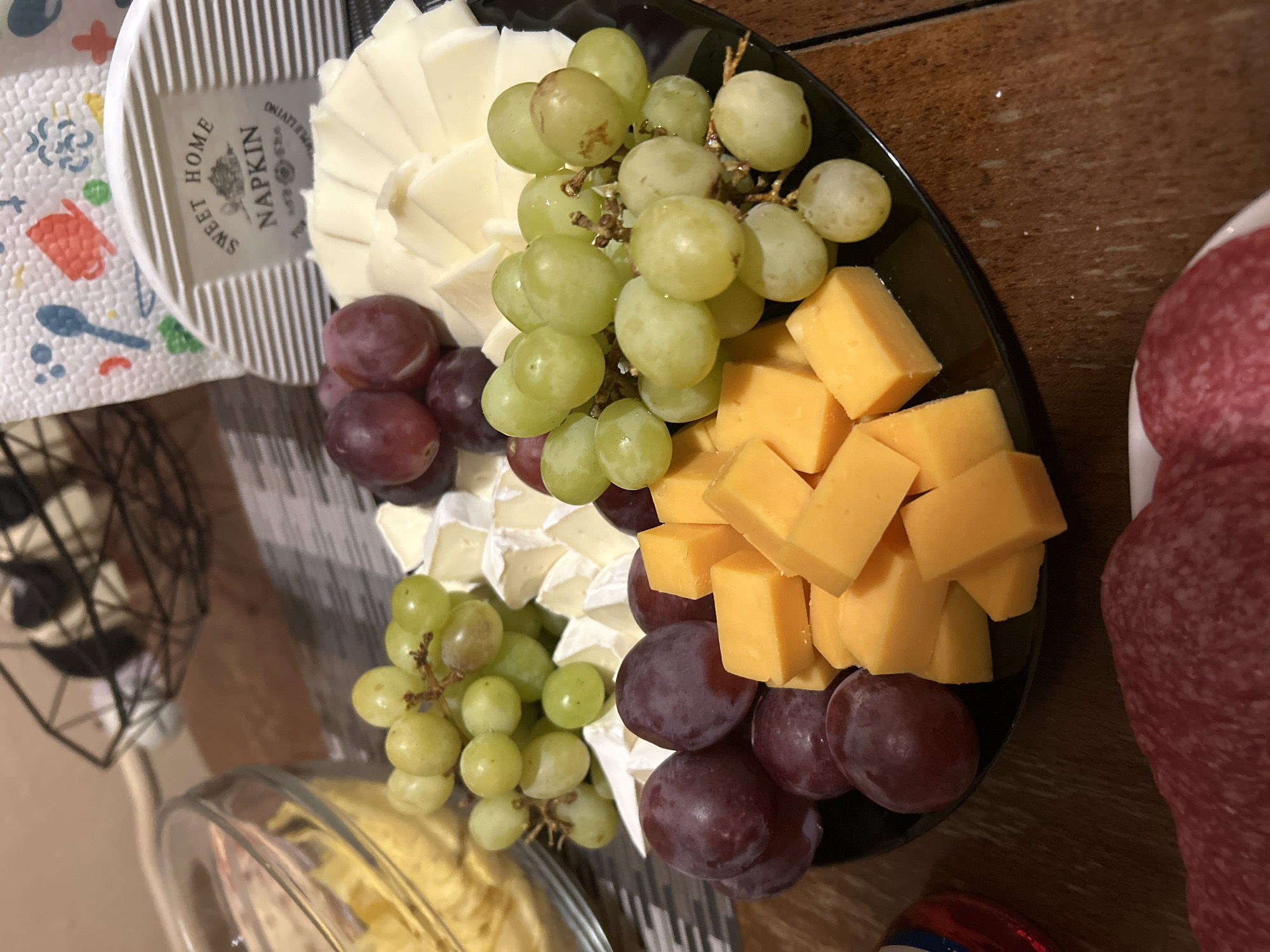 The image captures a close-up, top-down view of a charcuterie-style food spread arranged on a dark wooden table. An oval black serving platter dominates the scene, filled with a variety of cheeses and grapes.
On the platter, fan-shaped slices of white cheese are artfully fanned out alongside wedges of a soft, white-rinded cheese, possibly brie. Numerous cubes of bright orange cheddar cheese are piled in another section. Generous clusters of both green and red grapes are interspersed among the cheeses, adding vibrant color. To the right, a white plate holds slices of red cured meat, likely salami, partially visible. Below the platter, a clear glass bowl contains a yellowish substance, possibly a dip or a soft cheese spread.
To the left of the main platter, a white, ribbed, semi-circular object, likely a coaster or a small dish, rests on a patterned white napkin. The napkin features colorful, whimsical illustrations of kitchen items such as a red pot and a blue spoon. Text visible on the white ribbed object clearly reads "SWEET HOME NAPKIN" with a small emblem and "ONLIVING" printed below. A blurred, dark, geometric wire-frame object is partially visible in the bottom left corner.
The setup suggests a social gathering, party, or casual meal. The lighting appears to be artificial, indicating an indoor setting. No specific time of day is discernible, and while the context mentions Sofia, Bulgaria, there are no visual cues within the image to confirm this location. The overall presentation implies a pleasant and inviting atmosphere for snacking.