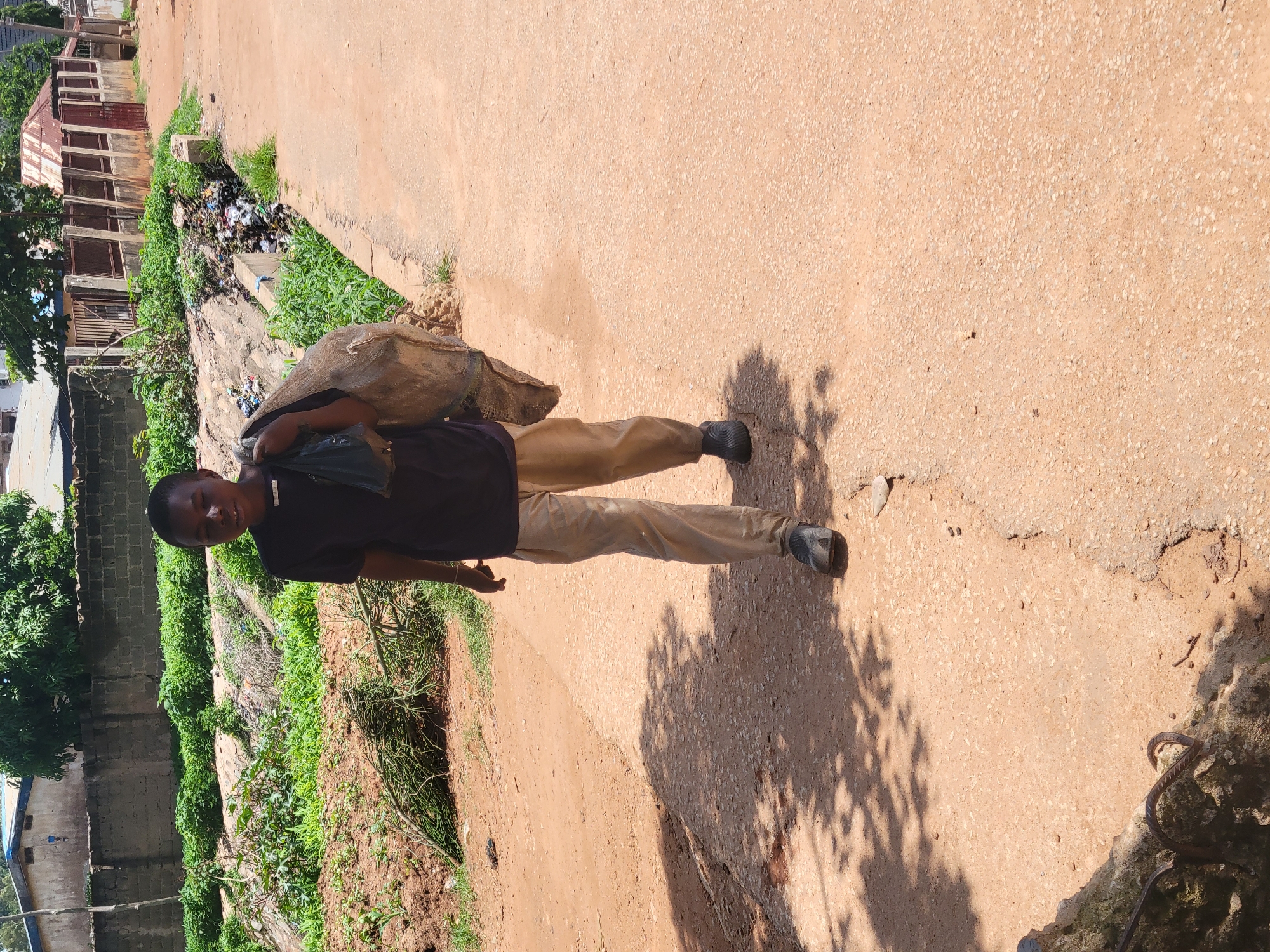 A young boy, appearing to be of Nigerian descent and around 10-12 years old, walks along a dirt road in Bukuru, Nigeria. He is carrying a large, rough-textured sack over his right shoulder, the contents of which are not clearly visible. The boy is wearing a dark t-shirt and light-colored trousers, with dark, utilitarian shoes. He is walking towards the viewer at a slight angle, his body positioned in the center of the frame. The road itself is unpaved, with a rough, sandy texture and evidence of cracking. Shadows of trees are cast across the road, indicating sunlight.
To the left of the road, there is a raised embankment covered in sparse vegetation and some scattered branches. Beyond this, a low concrete wall separates the road area from what appears to be residential or commercial buildings in the background. These structures have various roofing materials, including corrugated metal. A visible ditch runs along the side of the road further back, filled with a significant amount of debris, including plastic waste and other refuse, which is a prominent feature of the environment. The overall impression is one of a rural or semi-urban setting within Bukuru, Nigeria, characterized by a humble environment and the presence of litter.