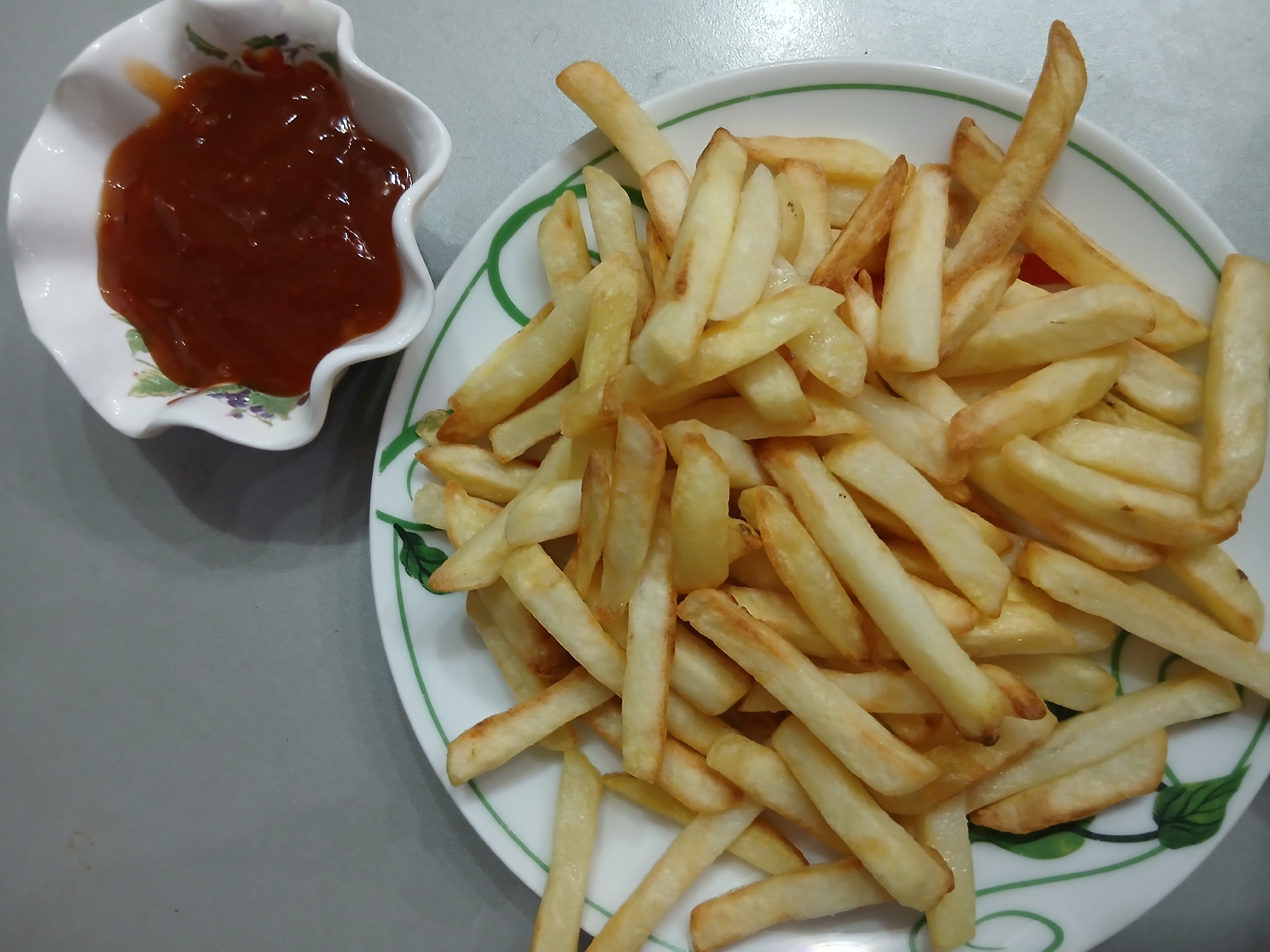 A plate of french fries is shown, along with a small bowl of ketchup. The french fries are golden brown and appear to be freshly fried. They are arranged in a pile on a white plate with a green and white decorative border. The ketchup is a deep red color and is in a white, scalloped bowl. The scene is set on a plain gray surface. There are no people or animals visible in the image.