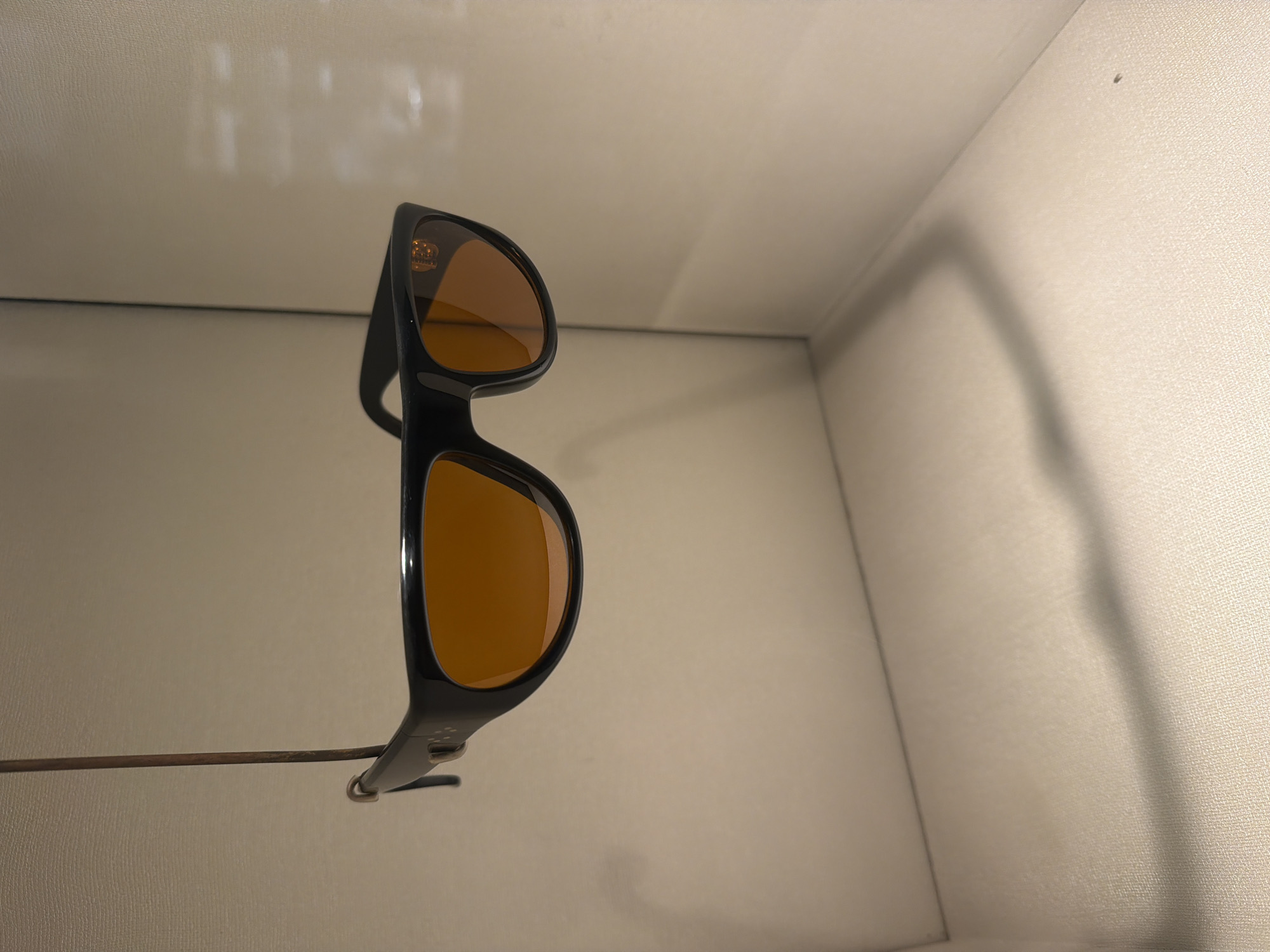 A pair of black-framed sunglasses with amber lenses are suspended on a thin metal rod in a corner, seemingly against a light beige or off-white textured background. The angle of the camera and the positioning of the sunglasses suggest they might be displayed in a store or exhibit. The scene gives the impression of a minimalist presentation, emphasizing the design of the sunglasses. Without further information, it is impossible to determine a specific location in Paris.
