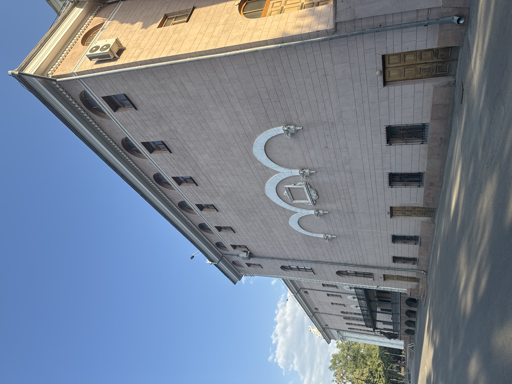 The image shows the exterior of a large, pink, neoclassical building. The building has a long facade with numerous windows, many of which are arched. There are also several decorative arches on the facade. The building appears to be old, with some signs of wear and tear. The sky is blue with some scattered clouds. Trees and a balcony are visible on the left side of the building. The ground in front of the building is paved and has shadows cast on it, indicating it is daytime. An air conditioning unit is mounted on the right side of the building. The building is likely a public institution or a historical landmark.