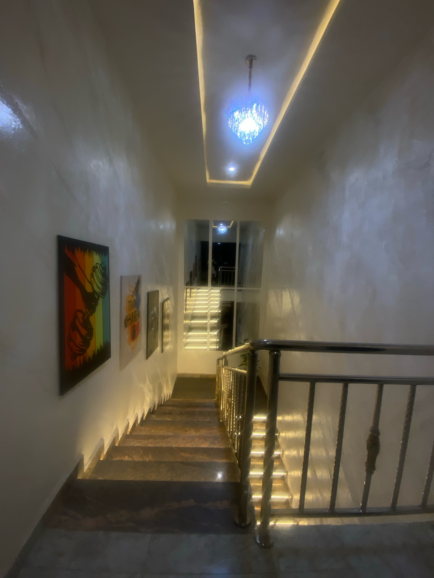 A well-lit interior hallway with a staircase in Barden Yana, Nigeria, features a modern chandelier at the ceiling and recessed lighting along the walls. The walls are textured and painted white. On the left wall, there are three framed pieces of art: the first depicts a dumbbell with a rainbow background, followed by two smaller, less distinct framed artworks. The staircase, made of polished granite, is illuminated from below by lights integrated into the risers, creating a dramatic effect. A stainless steel handrail lines the right side of the staircase. The environment appears to be a residential or commercial building, possibly a home or a hotel, designed with a contemporary aesthetic. There are no people or animals present in the image.