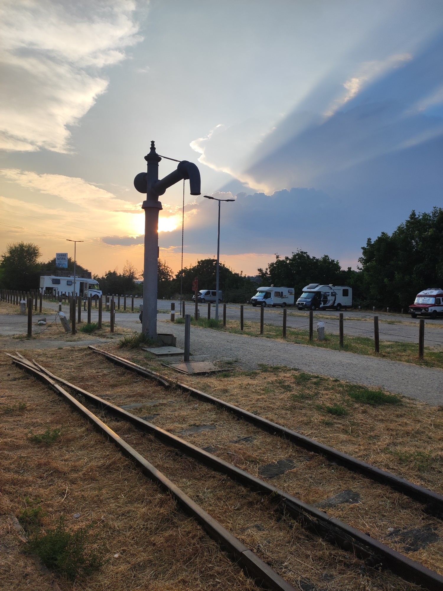 The image depicts an outdoor scene at sunset, with the sun casting long shadows and illuminating the sky with warm hues. In the foreground, a set of disused railway tracks run diagonally across the frame, surrounded by dry, yellowed grass.
The midground features a large, old-fashioned water pump, its metallic structure silhouetted against the sky. Behind the water pump, a gravel-covered parking area is visible, marked by wooden posts. Several recreational vehicles (RVs) are parked in this area, suggesting a campground or rest stop. A sign reading "CAMINO NATURAL" is visible on a bush in the distance.
The background is dominated by trees and the sunset sky, characterized by dramatic cloud formations and sunbeams. The overall atmosphere is serene and twilight-like.
The image is not located in Béjar, Spain. The sign "CAMINO NATURAL" indicates a natural path, often found in rural or natural park areas. These paths are common throughout Spain, but this specific location cannot be identified as Béjar without further information.