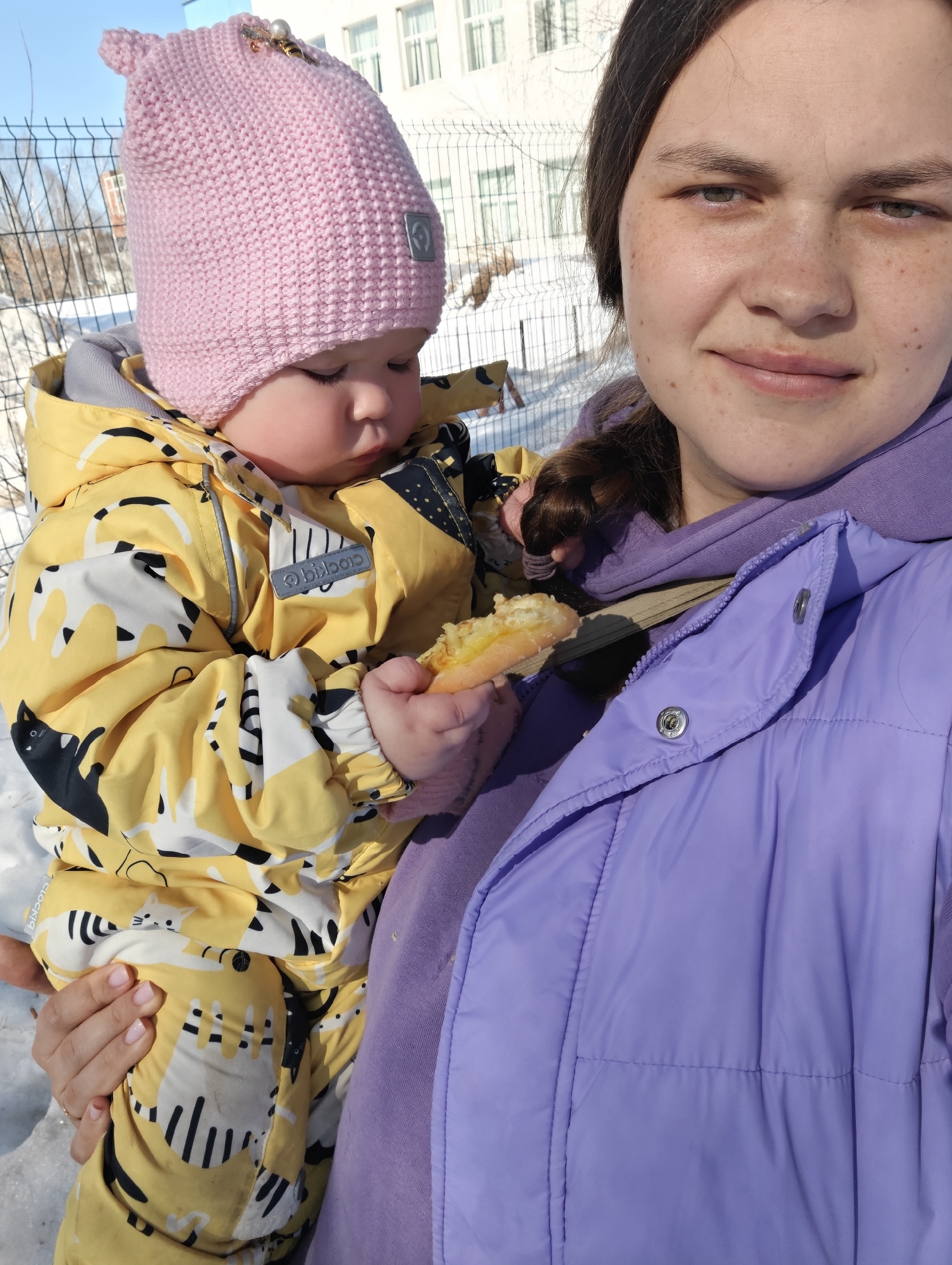 An outdoor scene in Chaikovsky, Russia, depicts an adult female holding an infant/toddler. The adult, positioned on the right, has brown hair and a freckled complexion, wearing a light purple hooded sweatshirt beneath a matching light purple puffer vest, and is looking towards the viewer with a slight smile. Her left hand, with light pink manicured nails and a gold-toned ring, supports the child. The child, positioned on the left, wears a yellow winter romper patterned with black and white cat motifs (brand "clocKid" visible) and a pink knitted beanie adorned with a pearl and metallic charm. The child holds a piece of pastry with a yellow filling in its right hand and is looking downwards. The ground is covered in snow. In the background, a green wire mesh fence and a white, multi-story building with multiple windows are visible under a clear blue sky.