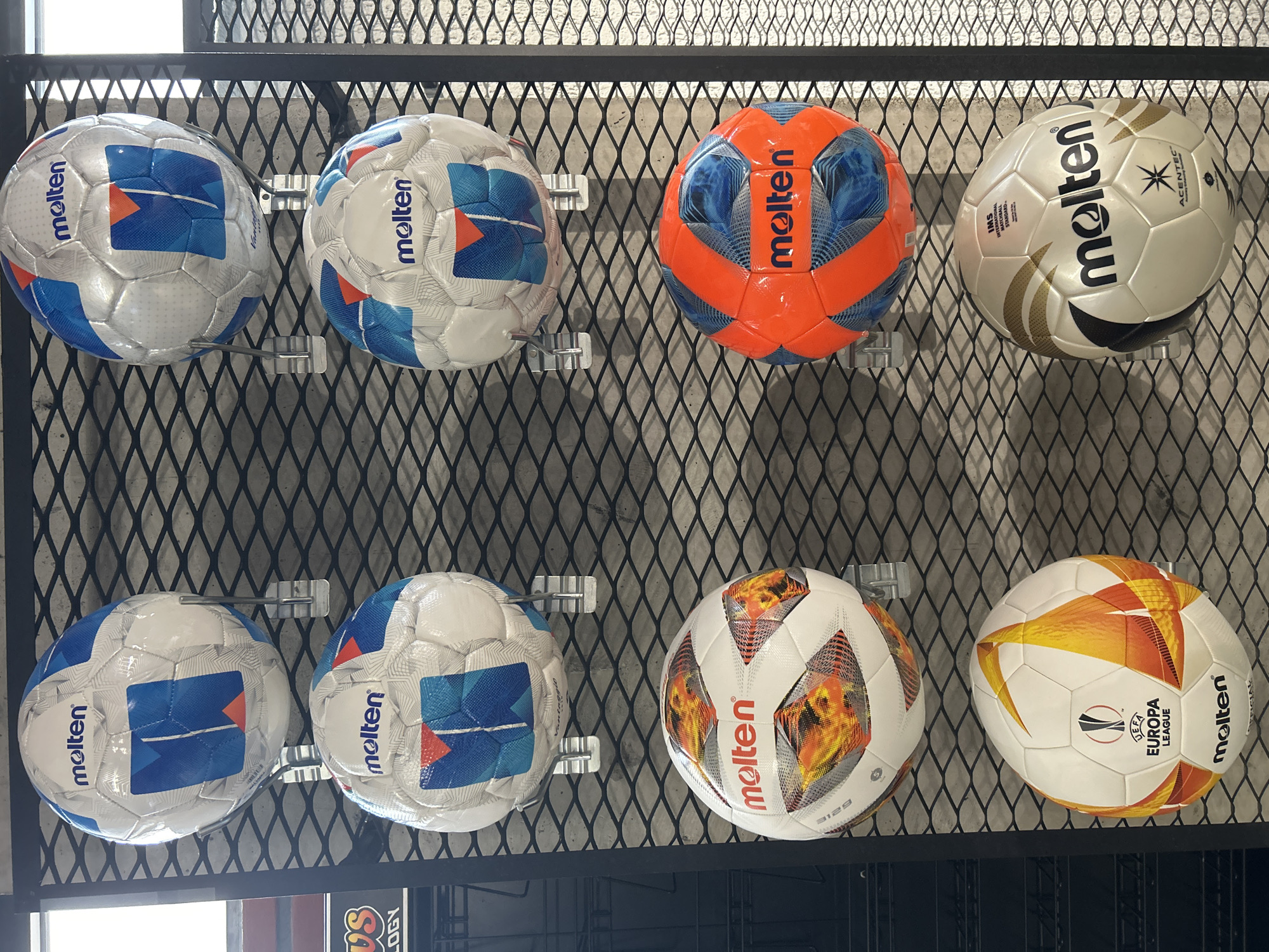 The image displays a display of eight Molten brand soccer balls, arranged in two rows of four, set against a diamond-patterned metal grid. The balls are of different designs. The environment suggests an indoor location, possibly a sporting goods store or a display area. The location is in the city of Tbilisi, Georgia, as indicated by the text below the display with a word containing "LOGY".