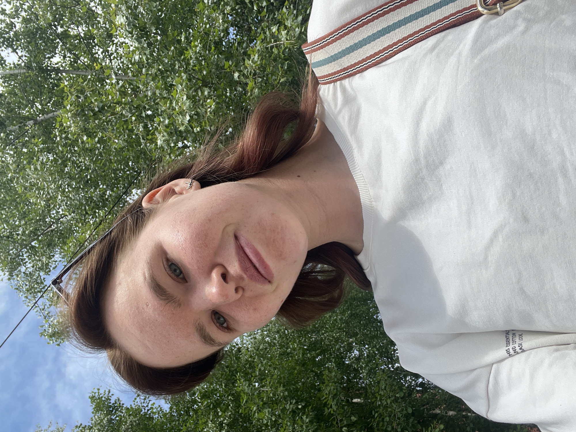 A selfie captures a woman with light skin, green eyes, and brown hair. She is wearing a white top with a striped strap visible. The background features green foliage, possibly trees. The angle suggests the picture was taken outdoors, looking up at the sky through the trees. While Chaikovsky, Russia is mentioned, there is no verifiable information in the image that confirms the location.