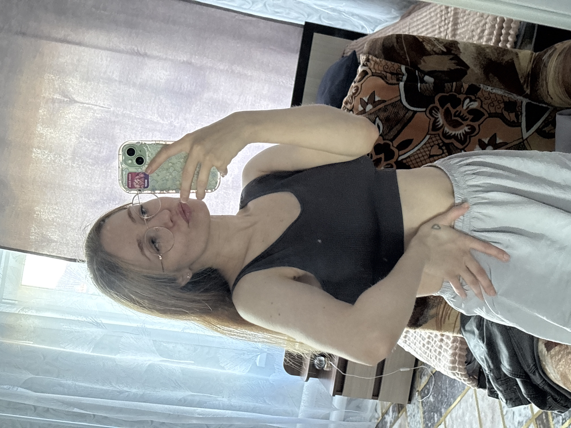 A young woman with blonde hair and glasses takes a selfie in what appears to be a bedroom. She is wearing a dark, ribbed crop top and light gray sweatpants. Her left arm is bent, holding a light green phone with a patterned case, and her right hand is resting on her sweatpants. A small heart tattoo is visible on the back of her left hand. Behind her, a patterned blanket or throw is draped over furniture, and sheer curtains hang near a window, suggesting it is daytime. The room appears somewhat cluttered with clothing and other items. The woman's expression is neutral, and the overall mood is casual and relaxed. No visible text is present in the image.
