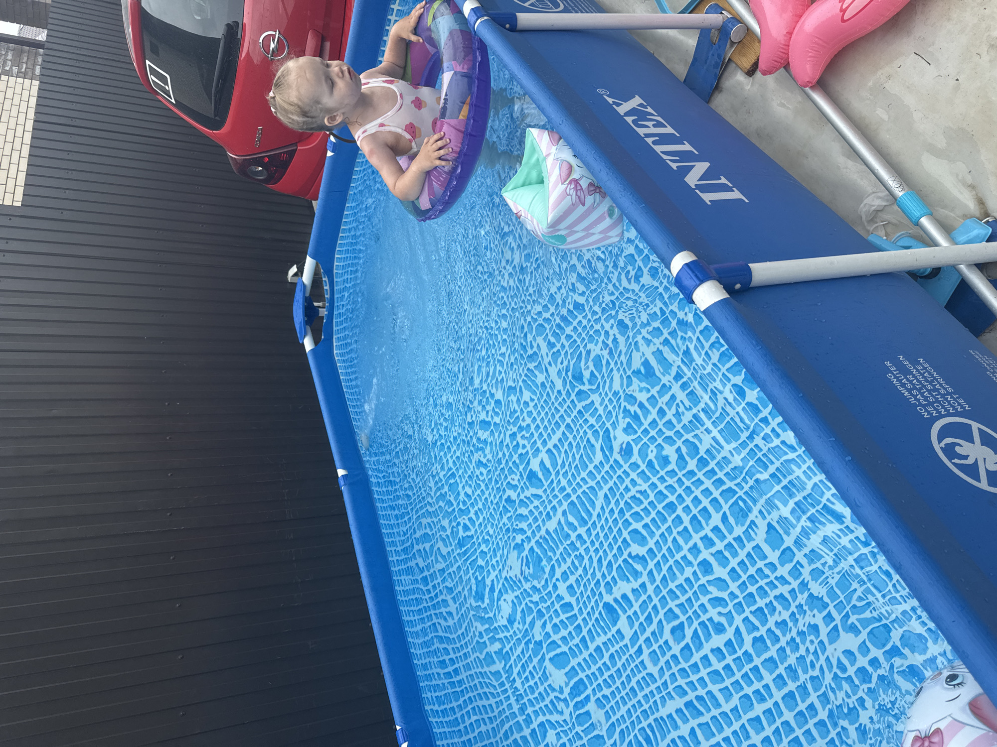 A young child wearing a swimsuit with a fruit pattern is sitting in a bright blue inflatable pool. The child is holding onto a colorful inflatable ring and has an arm float with a unicorn design on one arm. The pool is filled with clear water that has a grid pattern at the bottom. Next to the pool, a red car with an Opel logo is visible. The background shows a dark, corrugated metal fence and a brick wall. The scene appears to be set on a sunny day outdoors, possibly in a backyard. Text visible on the pool includes "INTEX" and warnings in multiple languages such as "NO JUMPING" and "NE PAS SAUTER".