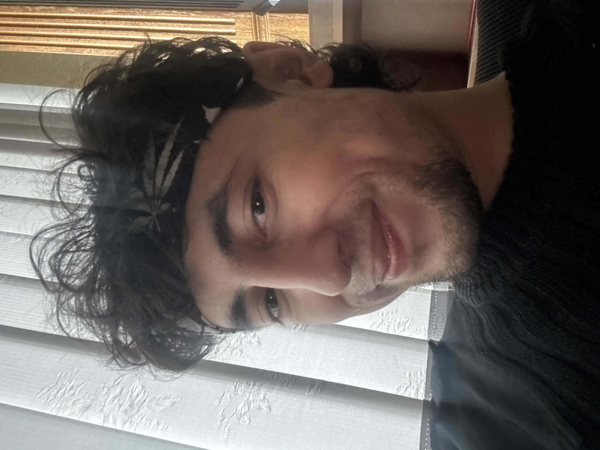 The media file is a portrait of a young man with dark, curly hair and a light complexion. He is wearing a black top or sweater, and a black bandana with a white pattern around his forehead. The man is smiling and looking towards the camera, with his head tilted to the side. His right eye is clearly visible, while his left eye is partially obscured by his hair. He has stubble on his chin and jawline.
The background consists of white blinds with a textured pattern, and a golden-colored object at the top of the frame, possibly a shelf or a part of the room's decor. The lighting suggests it might be daytime, with light filtering through the blinds. The overall mood appears casual and friendly, conveyed by the man's smile. The image is oriented vertically, as if taken by a phone. There is no visible text in the image.