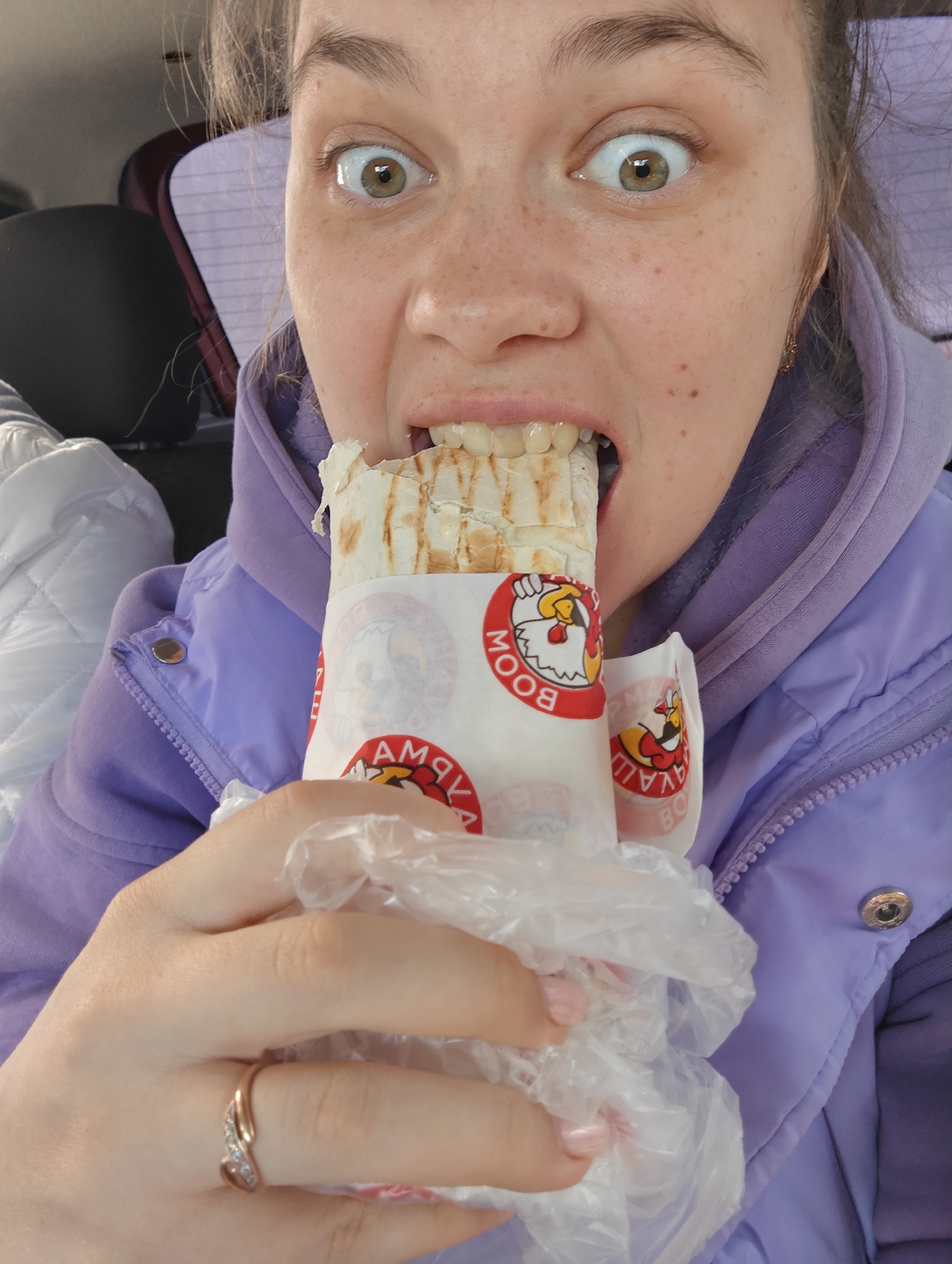 A female individual is depicted in a close-up shot, seated inside what appears to be a vehicle. She has light brown hair, green-brown eyes, and facial freckles. Her eyes are wide open, and her mouth is agape, biting into a lavash wrap or shawarma. She wears a purple hooded jacket over a lighter purple top. On her left ring finger, a gold-toned ring with small clear stones is visible. Her fingernails are painted light pink. The lavash wrap is partially enclosed in a white paper wrapper with repetitive red circular logos depicting a stylized red chicken head and the Cyrillic text "БУМ" (BOOM) and "АККА" (AKKA), identifying the "Akka Boom" brand. The wrap is also contained within a clear plastic bag. The background shows a dark car seat headrest to the left, a quilted white fabric object further left, and a faint purple patterned surface behind her head. The food item, an Akka Boom shawarma, indicates a likely purchase from an Akka Boom establishment in Chaikovsky, Russia, such as the one located at Проспект Победы, 30.