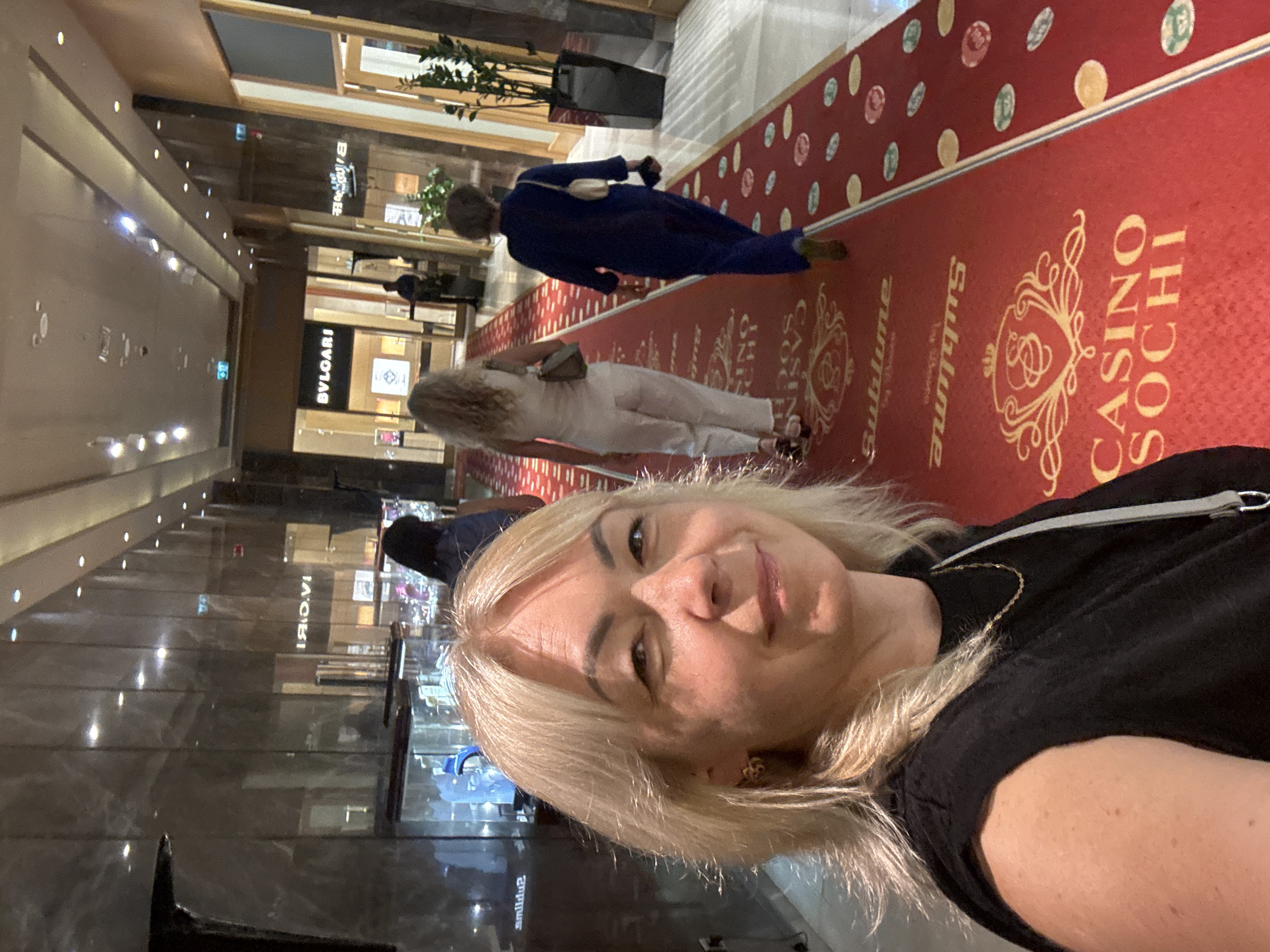 In Krasnaya Polyana, Russia, a woman with blonde hair and a black top takes a selfie in a casino hallway. She is in the foreground, smiling at the camera. Behind her, a red carpet leads down the hallway, adorned with various logos and designs including "CASINO SOCHI" and "Sublime". Three other women, dressed in casual attire, are walking away from the camera on the carpet. The hallway features mirrored walls, decorative lighting, and storefronts, one of which displays the "BVLGARI" logo. The environment appears to be an upscale shopping or entertainment area within the casino.