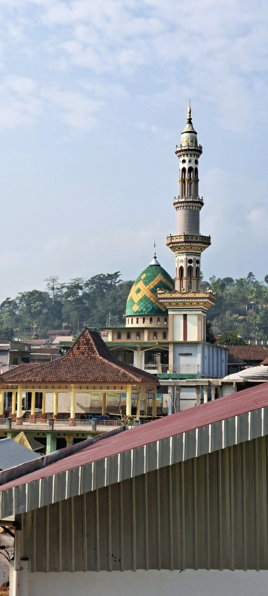 This is a vertical, eye-level, medium shot photograph taken on a clear day in Duyung, Indonesia. The image focuses on a mosque complex situated on a hillside. The most prominent structure is a tall, slender minaret with intricate detailing, topped by a spire. Adjacent to the minaret is the mosque itself, which features a distinctive green and yellow tiled dome. In the foreground, a large, modern building with a corrugated metal roof partially obscures the view of other structures within the complex. To the left of the mosque, a building with a brown, tiled, pyramidal roof and open-air verandas supported by yellow pillars is visible. The background is dominated by a densely forested hillside, indicating a semi-rural or suburban setting. The sky is a pale blue with scattered white clouds. There are no people visible in the image.