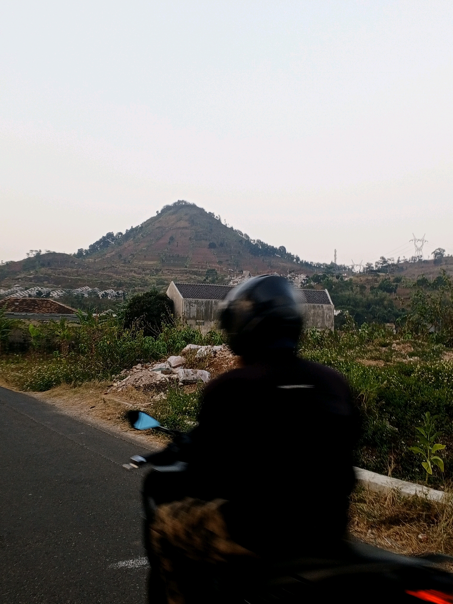 An over-the-shoulder shot shows a person wearing a black shirt and helmet, blurred due to motion, riding a motorcycle on an asphalt road. The road runs alongside a grassy embankment with scattered white bags and some foliage. In the background, a cone-shaped mountain dominates the landscape, with houses and vegetation dotting its slopes. A few buildings, including one with a gray concrete exterior and a tiled roof, are visible in the midground, nestled among trees. Power lines and transmission towers are also present on the hilly terrain in the distance. The sky is overcast and pale. The scene appears to be a rural or semi-rural area during the daytime.