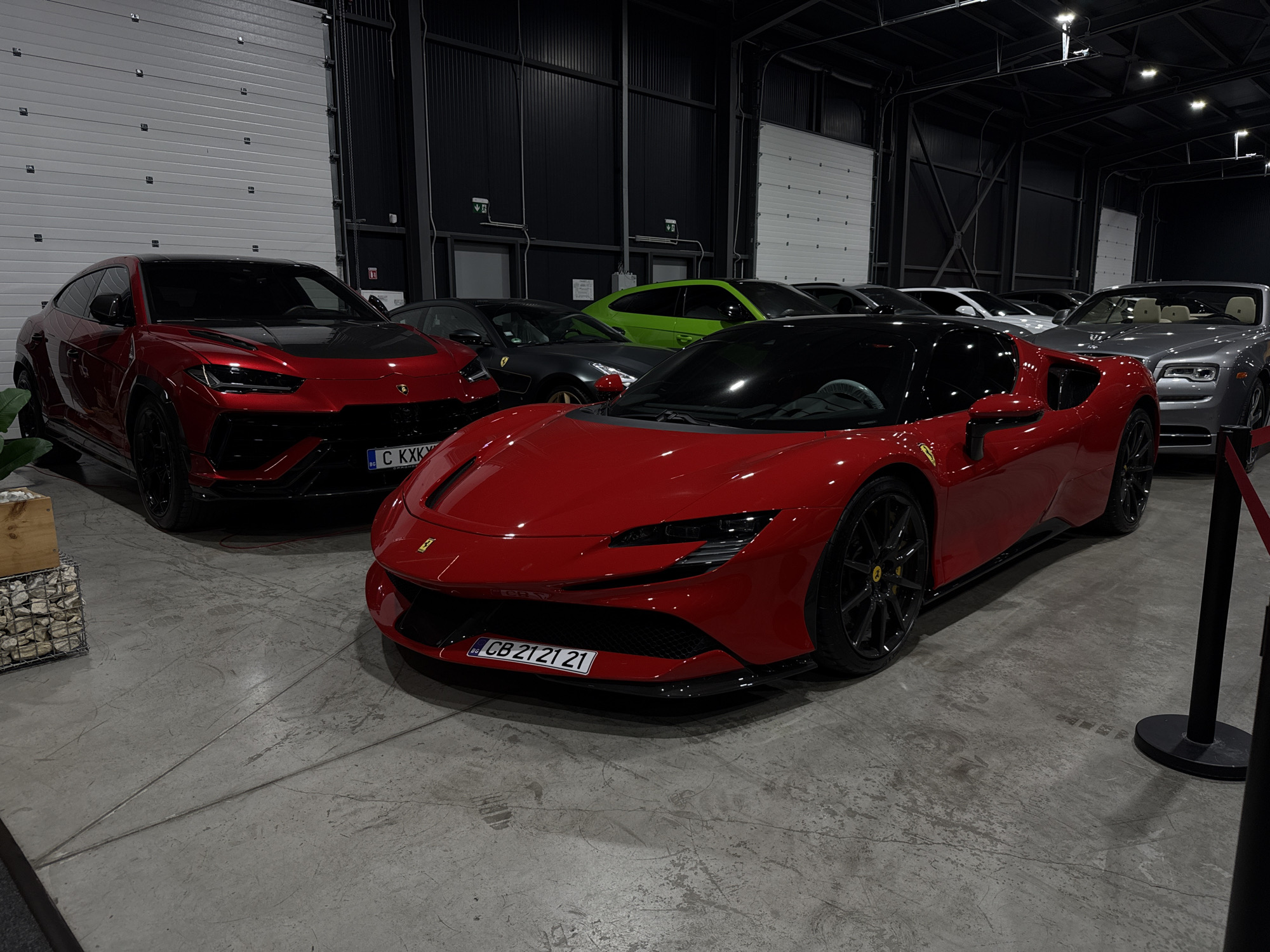 The image shows a collection of luxury cars displayed indoors, likely in a showroom or at an event. In the foreground, a bright red Ferrari is prominently featured, with its front bumper and license plate clearly visible. The license plate reads "CB 21 21 21". To the left of the Ferrari, a red Lamborghini SUV is parked, and behind it, other cars including a green Porsche, a dark grey car, and a silver Rolls-Royce are visible. The environment appears to be a large, open space with industrial-style walls and a concrete floor. The lighting suggests an indoor setting with artificial lights overhead. The overall scene conveys a sense of wealth and automotive passion. The location is identified as being in Plovdiv, Bulgaria, suggesting this display is part of a local car show, dealership event, or private collection event within the city.