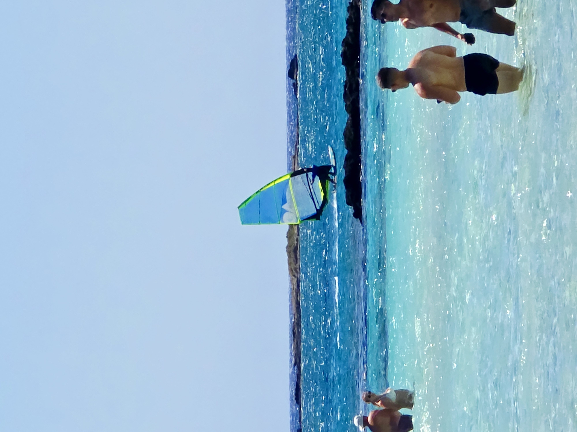 In an outdoor, daytime shot, two men stand in shallow, clear turquoise water, facing away from the camera, with a third man and a woman standing in the water to their left, also facing away. The man on the left wears a white hat and dark swim trunks, while the woman wears a white bikini. A windsurfer with a blue and green sail navigates the choppy, blue ocean in the middle ground, with dark rock formations dotting the sea. In the distance, a rocky shoreline is visible against a pale blue sky.
