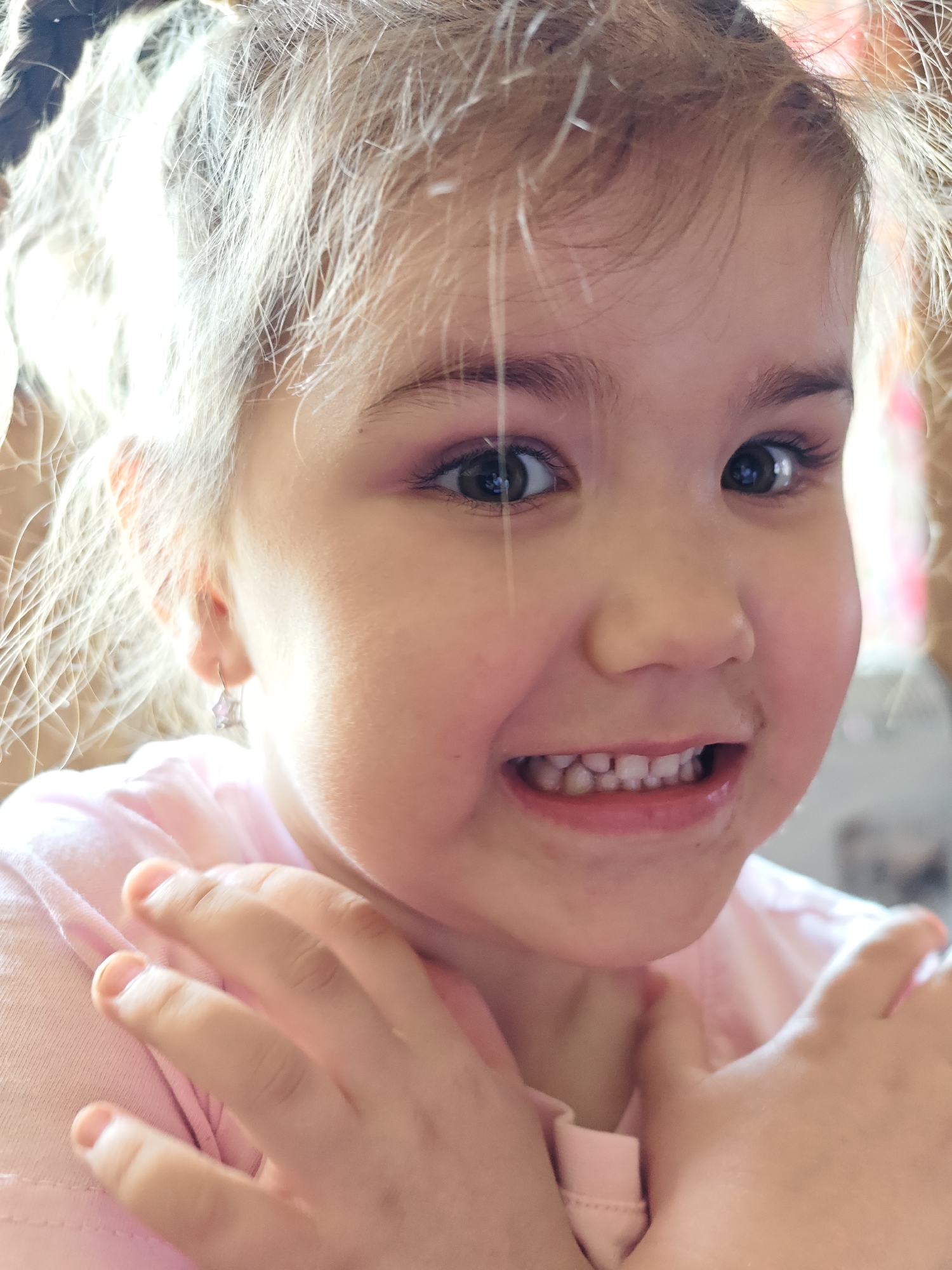 A close-up shot depicts a young girl with fair hair, styled in pigtails. She is wearing a light pink shirt. The girl is smiling, revealing her teeth, and has dark smudges around her mouth. Her hands are raised near her chest, with her fingers spread. The girl is wearing a star-shaped earring in her left ear. The location is not discernable as Malyi Bukor, Russia.