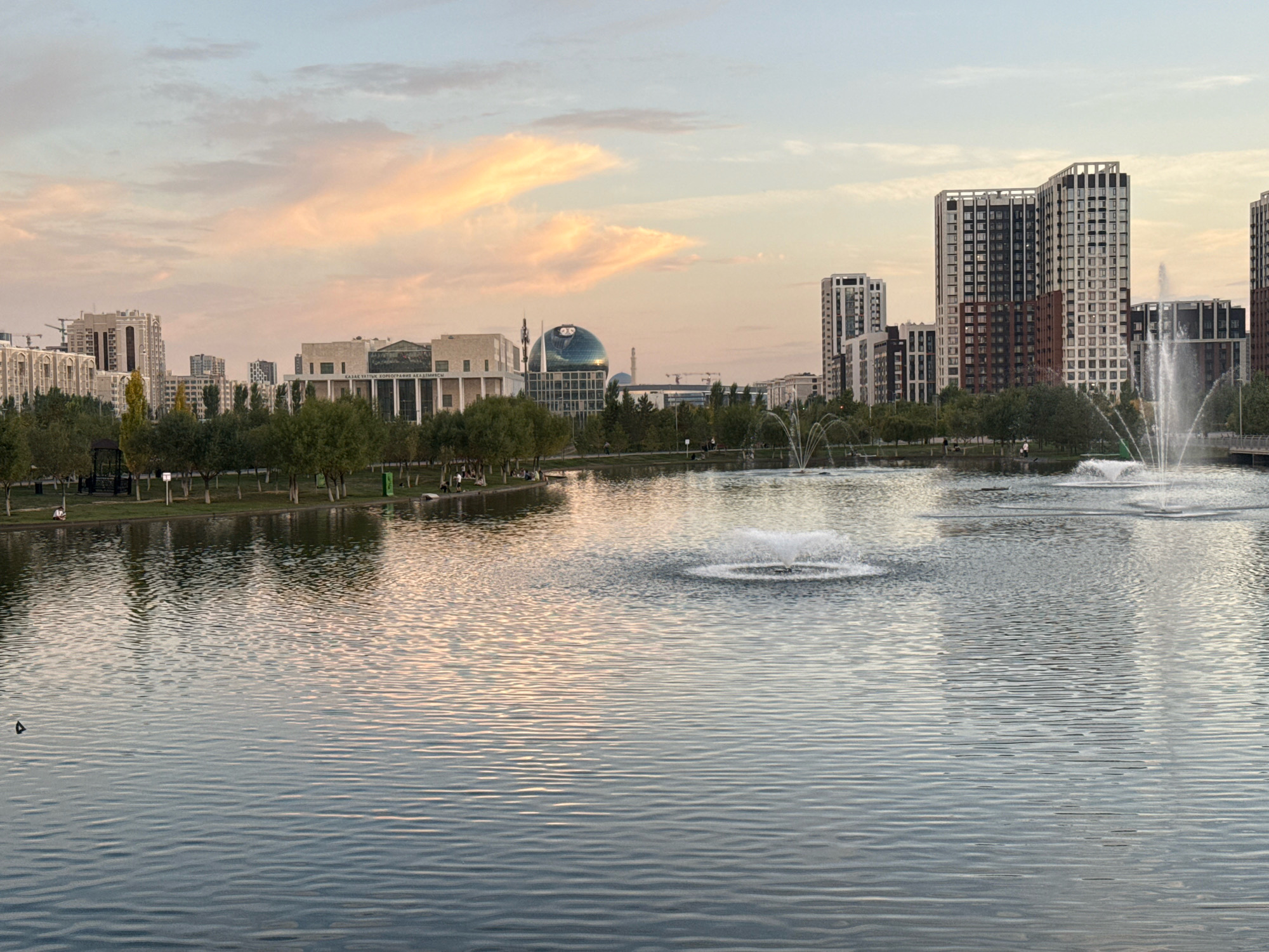 The image depicts a serene lake in Astana, Kazakhstan, at dusk. The calm waters of the lake reflect the soft hues of the sunset, with streaks of pink and orange painting the sky. Several fountains are active in the lake, their plumes of water catching the light. In the background, modern high-rise buildings dominate the cityscape, interspersed with trees and greenery. One prominent structure is the Bayterek Tower, recognizable by its spherical glass top and spire. The scene suggests a peaceful urban park setting, likely a popular spot for relaxation and recreation. The overall atmosphere is tranquil and picturesque, capturing the beauty of Astana as day transitions into night.