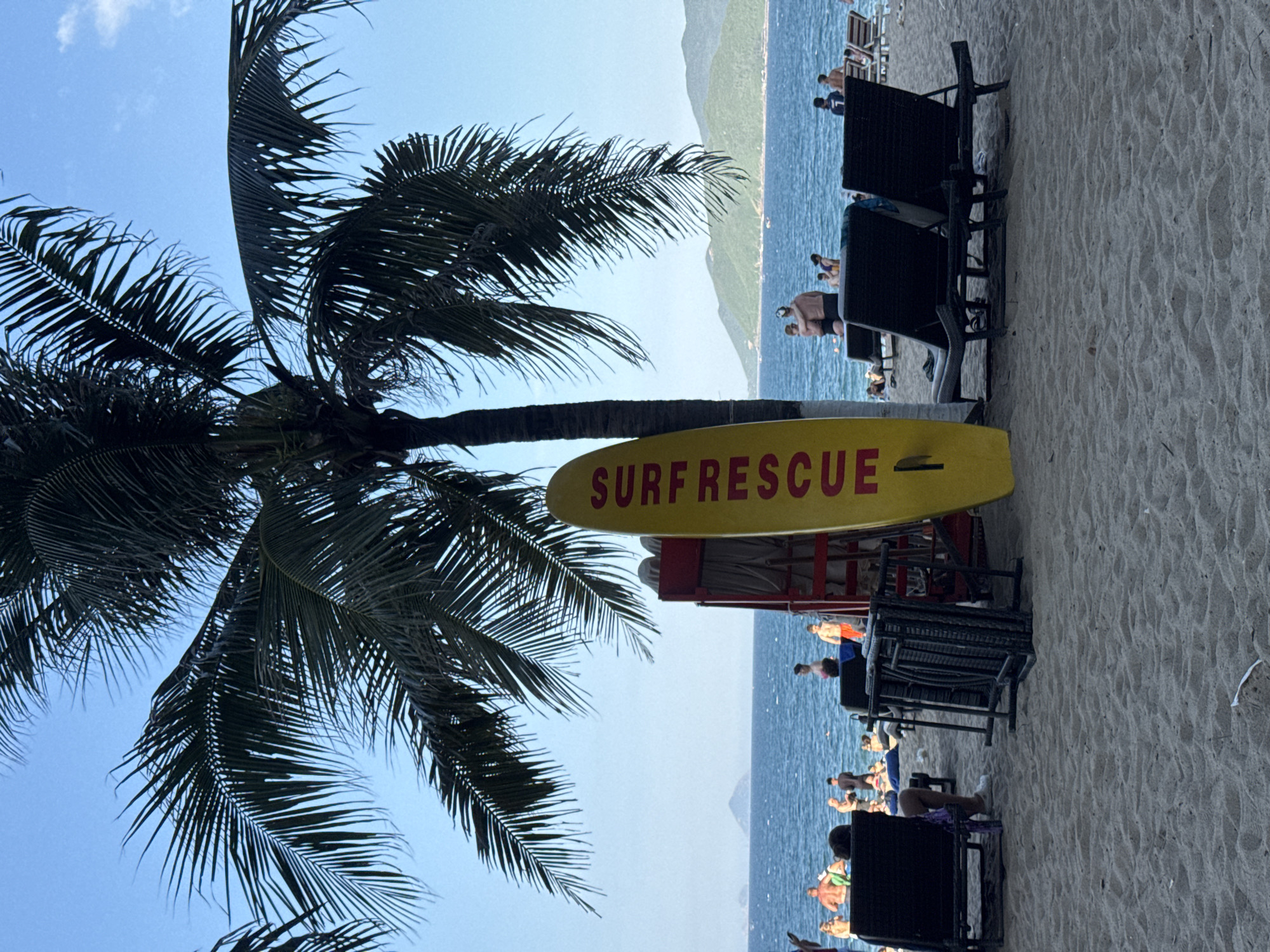 The image depicts a tropical beach scene in Nha Trang Ward, Vietnam. In the foreground, a bright yellow surfboard with red lettering spelling "SURF RESCUE" stands upright, leaning against a red lifeguard stand. Several black lounge chairs are arranged on the sandy beach. In the middle ground, the blue ocean stretches out, with a few people visible in the water and on the shoreline. The background features lush green mountains under a clear blue sky with scattered clouds. Palm trees are prominent in the upper portion of the image, their fronds casting shadows on the beach. The scene suggests a warm, sunny day at a popular beach destination.