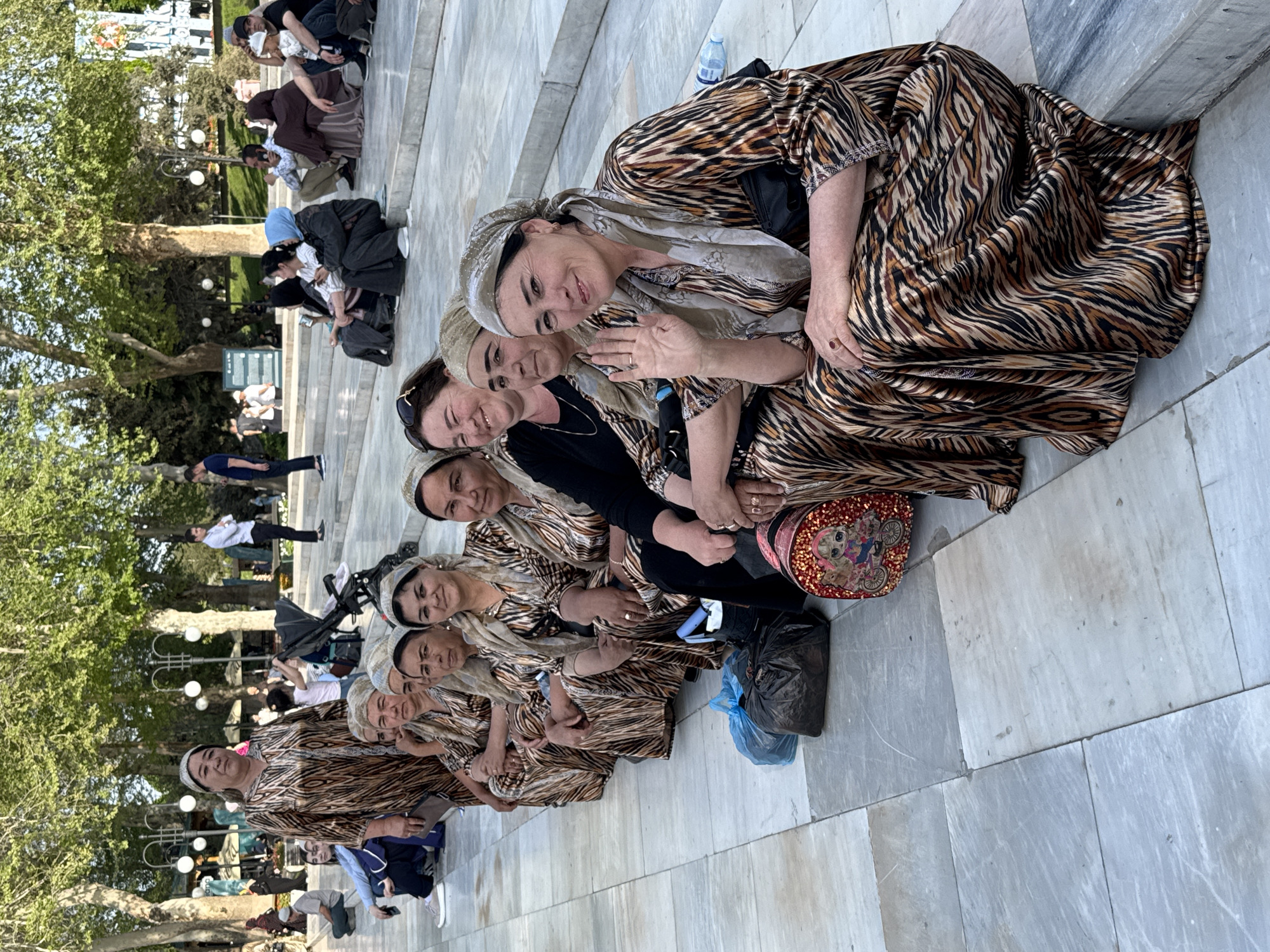 A group of women are seated on the stone steps in the city of Samarkand City, Uzbekistan. The women are wearing matching tiger-striped dresses and headscarves. They are smiling and looking at the camera. Behind them, there is a crowd of people, trees, and street lights. A woman’s purse with cartoon characters is visible to the right. A black and blue plastic bag is also visible to the right. A stroller is visible among the crowd.