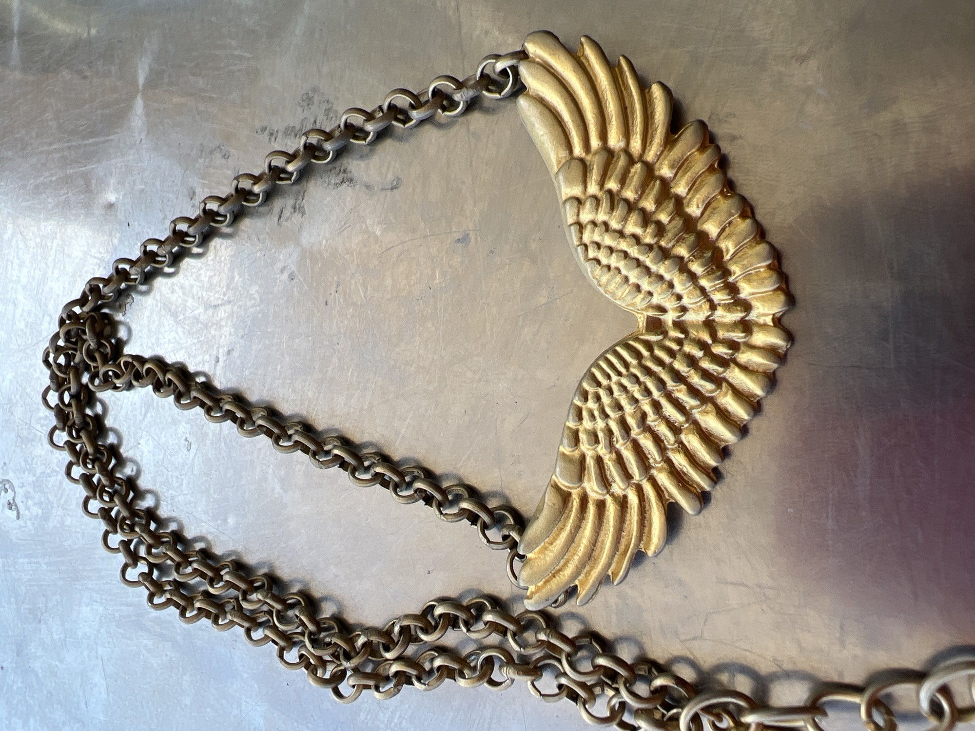 The image depicts a gold-colored metal necklace with a stylized wing pendant lying on a brushed metal surface. The necklace is made of interlocking circular links. The pendant consists of two wings, densely detailed with individual feathers. The background surface is slightly reflective, showing subtle gradients in color and texture. There is no evidence to suggest that the photograph was taken in Baošići, Montenegro.