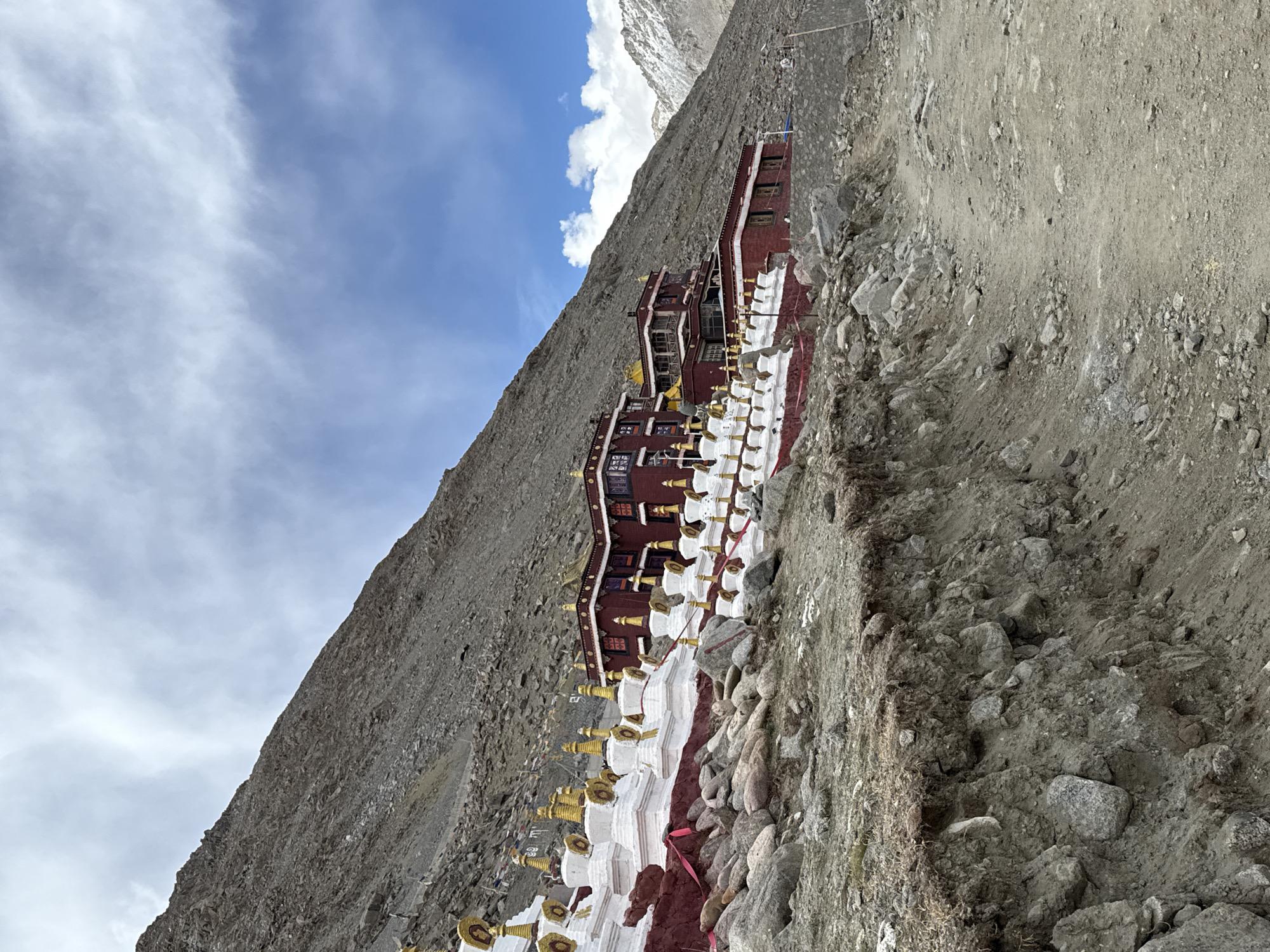 The image shows a multi-story, maroon-colored building nestled on a steep, rocky hillside in China. The building has visible windows and a golden decorative roof. A row of white stupas with golden tops extends from the base of the building, creating a visual path down the hill. The sky is partly cloudy. The ground is rocky and uneven. The building is likely a Buddhist temple or monastery. There are no people visible. The location is likely a mountainous region, potentially in the Parga town area of China.