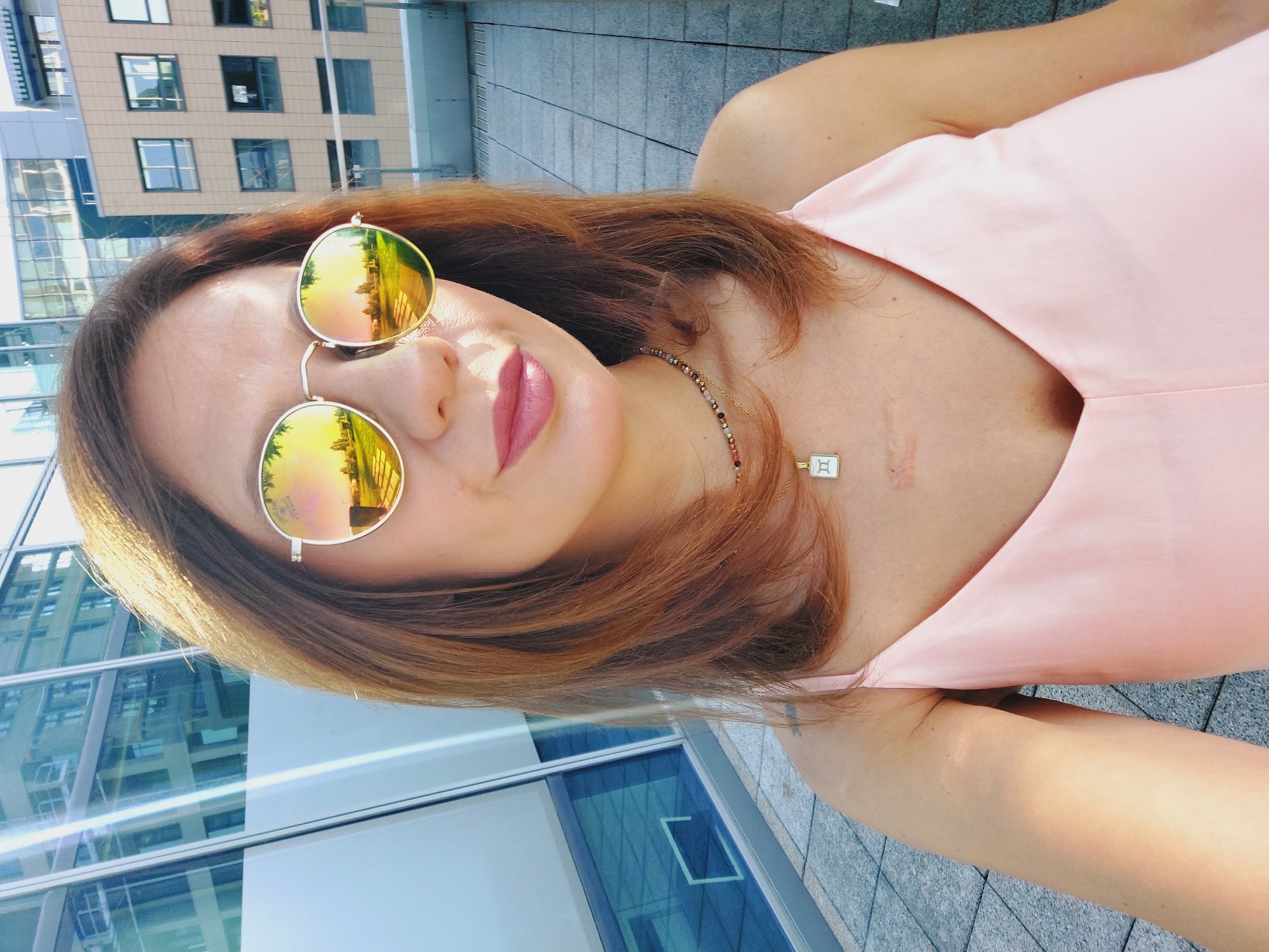 An outdoor, medium-shot selfie captures a woman with reddish-brown hair and fair skin, wearing round, mirrored sunglasses with yellow-gold lenses that reflect a cityscape and grassy area. She is wearing a light pink, spaghetti-strap top and a beaded necklace with a white pendant that features a Gemini symbol. A visible scar is present on her chest. She is standing on a tiled patio, with a modern building featuring large glass windows and another building with tan siding visible in the background. The lighting suggests it is daytime.